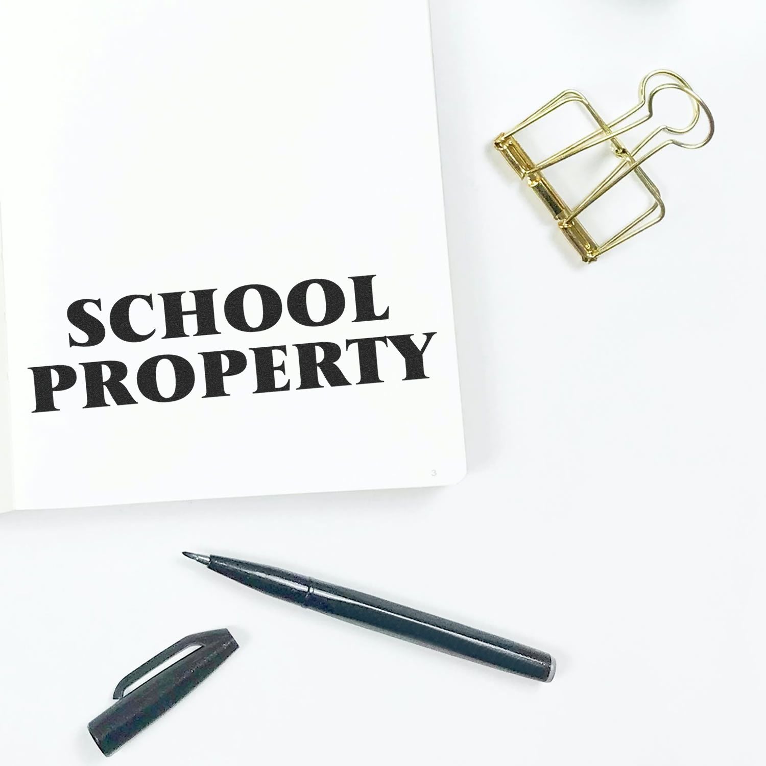 Self Inking School Property Stamp marking SCHOOL PROPERTY on paper, with a pen, cap, and gold binder clip nearby on a white surface.