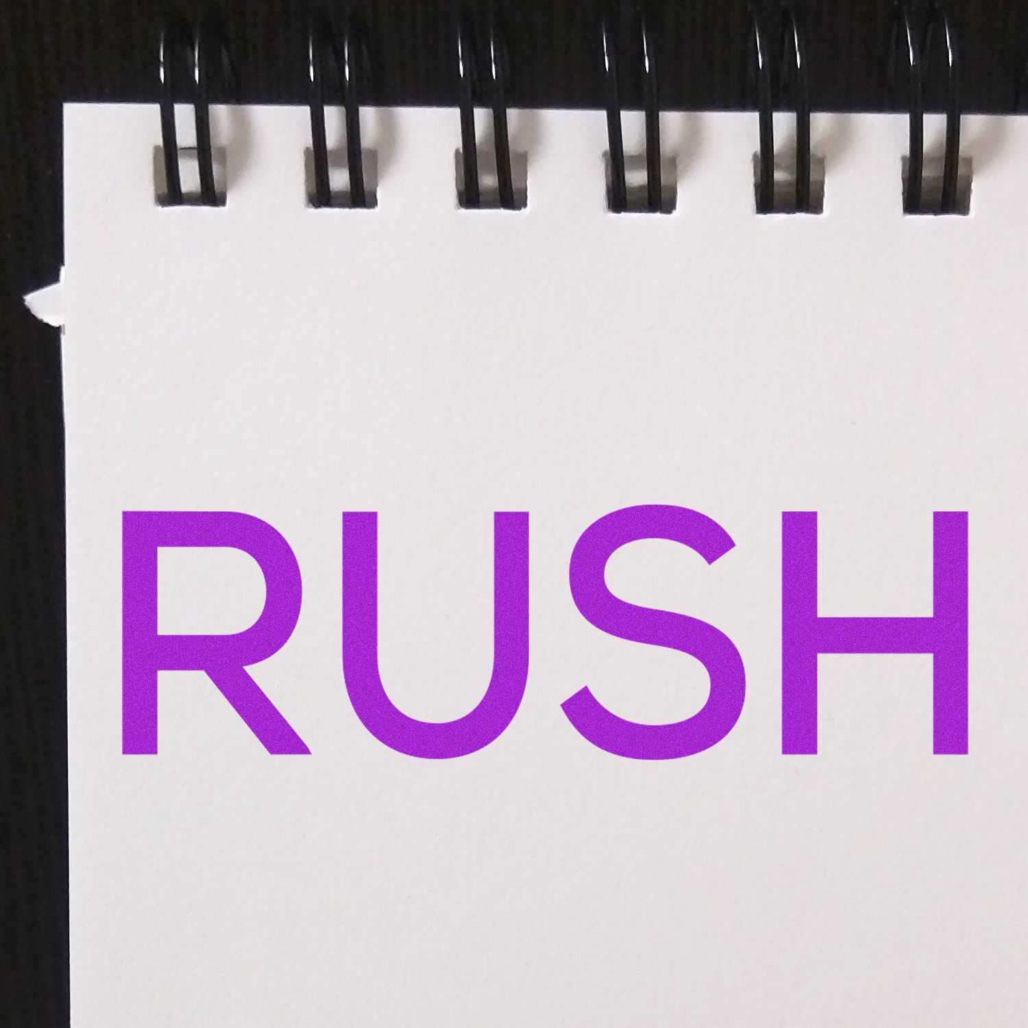 A Slim Pre-Inked Skinny Rush Stamp imprinting the word RUSH in bold purple letters on a white notepad with black rings.