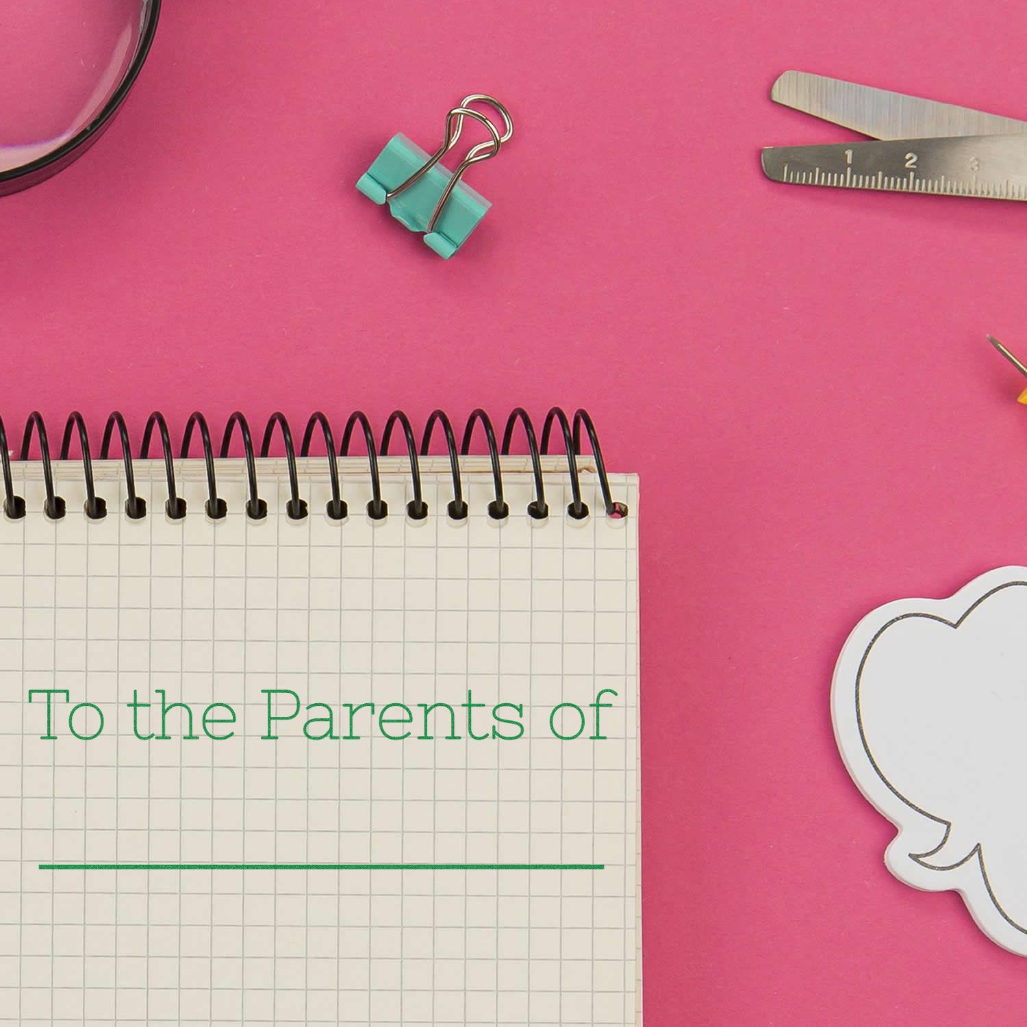 Self Inking Skinny To the Parents of Stamp in use on a notebook, surrounded by office supplies on a pink background.