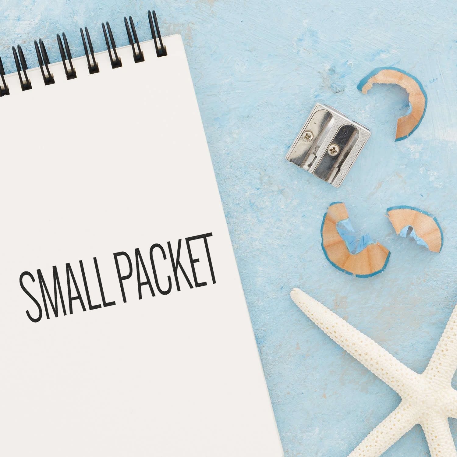 A notebook with Small Packet text, a pencil sharpener, shavings, and a starfish on a blue surface. Large Small Packet Rubber Stamp.