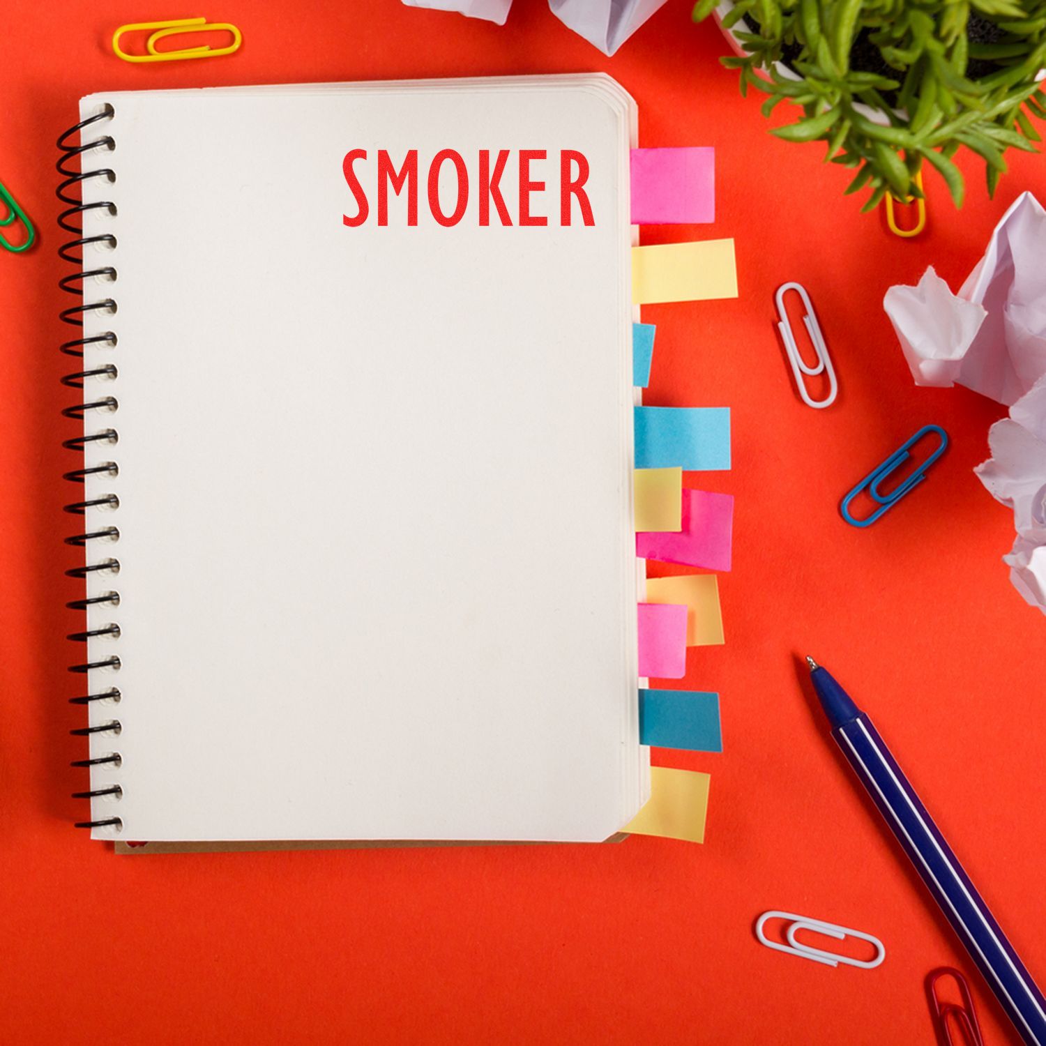 Notebook stamped with SMOKER using the Self Inking Smoker Stamp, surrounded by colorful paper clips, sticky notes, and a pen on a red background.