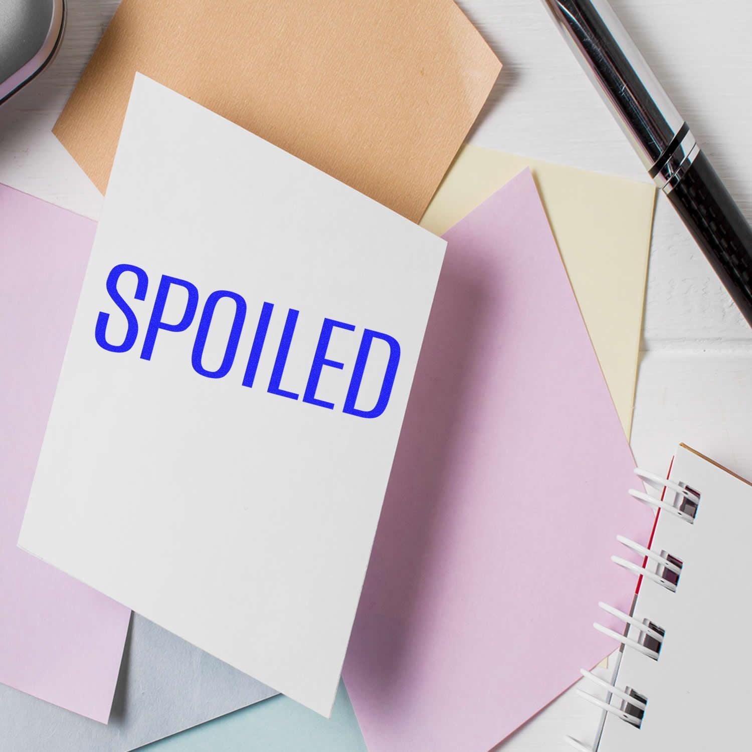 Large Spoiled Rubber Stamp in blue ink on a white card, surrounded by pastel-colored papers and a pen on a white desk.