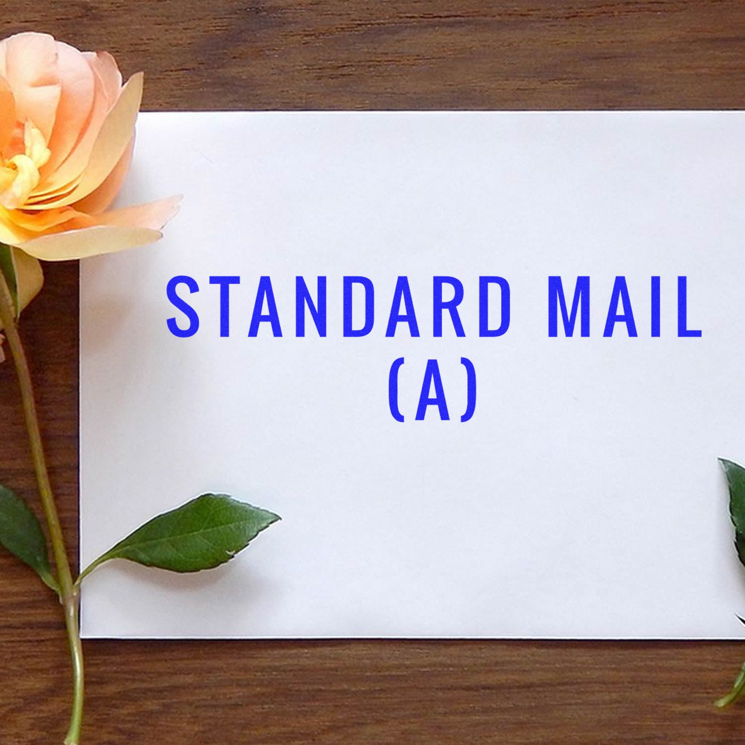 Envelope stamped with STANDARD MAIL (A) in blue ink using the Large Self Inking Standard Mail (A) Stamp, placed on a wooden surface with flowers.