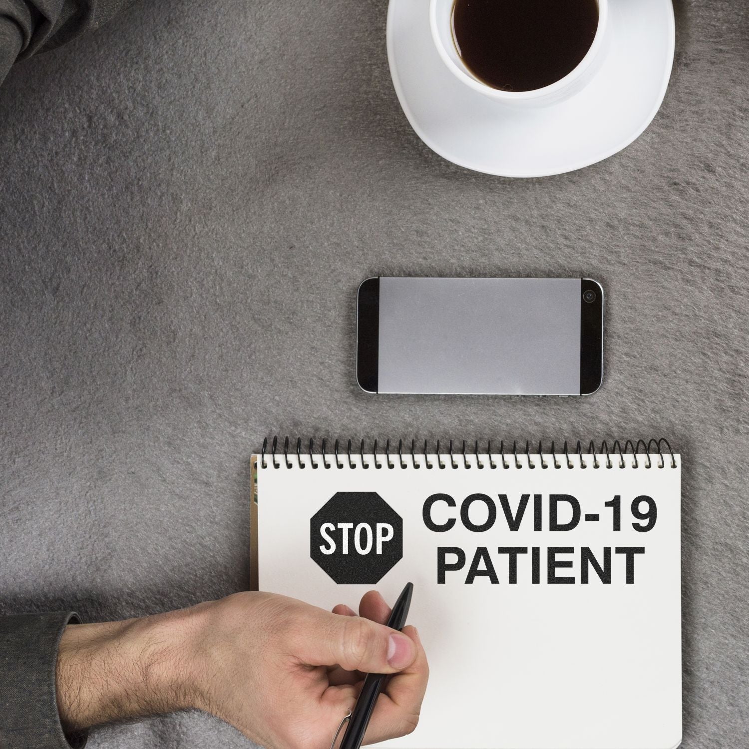 Hand holding pen near notebook with 'Stop Covid-19 Patient' rubber stamp, smartphone, and coffee cup on table.