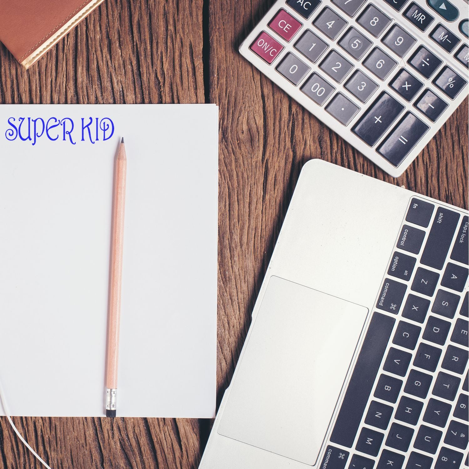 Large Super Kid Rubber Stamp on paper beside a pencil, laptop, calculator, and notebook on a wooden desk.