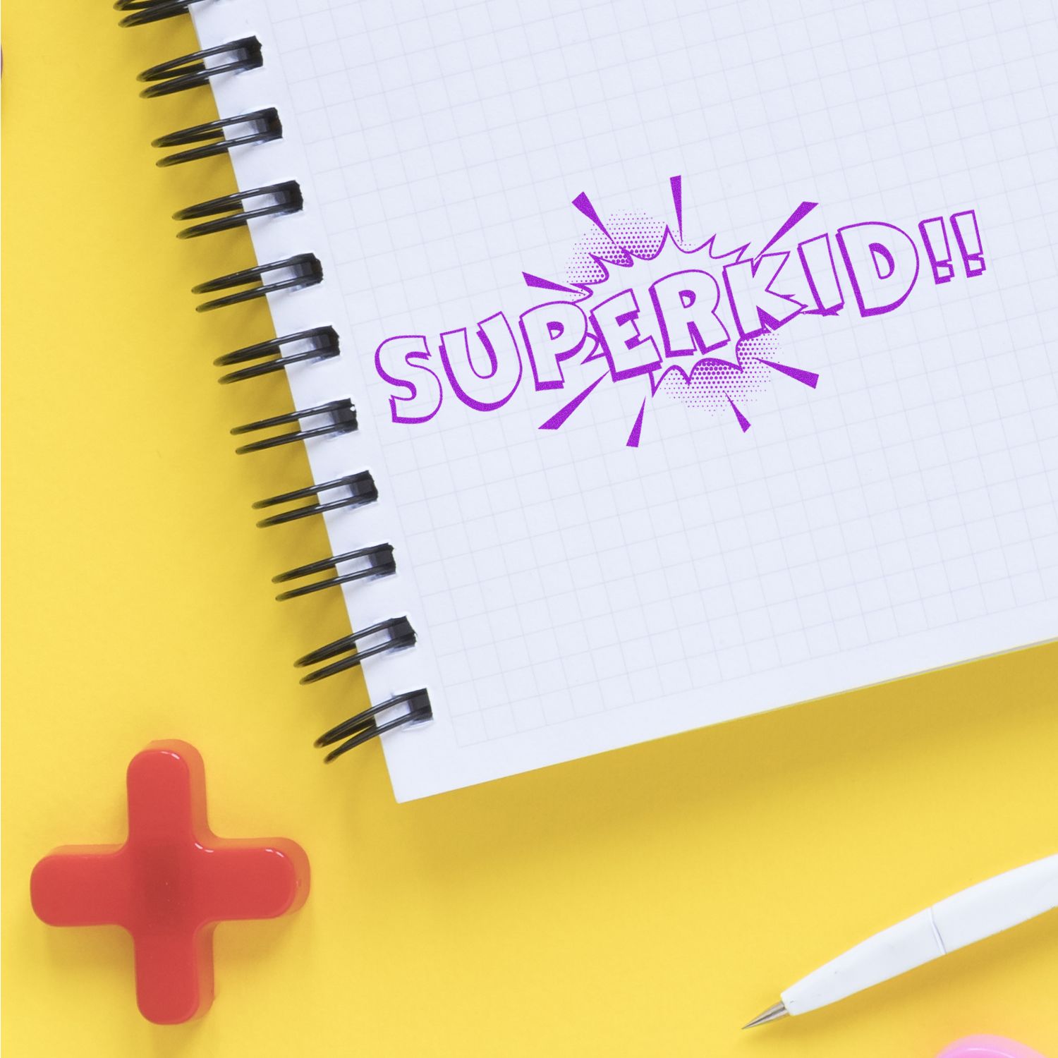 Large Superkid Rubber Stamp imprint on a spiral notebook with a yellow background, red cross, and white pen nearby.