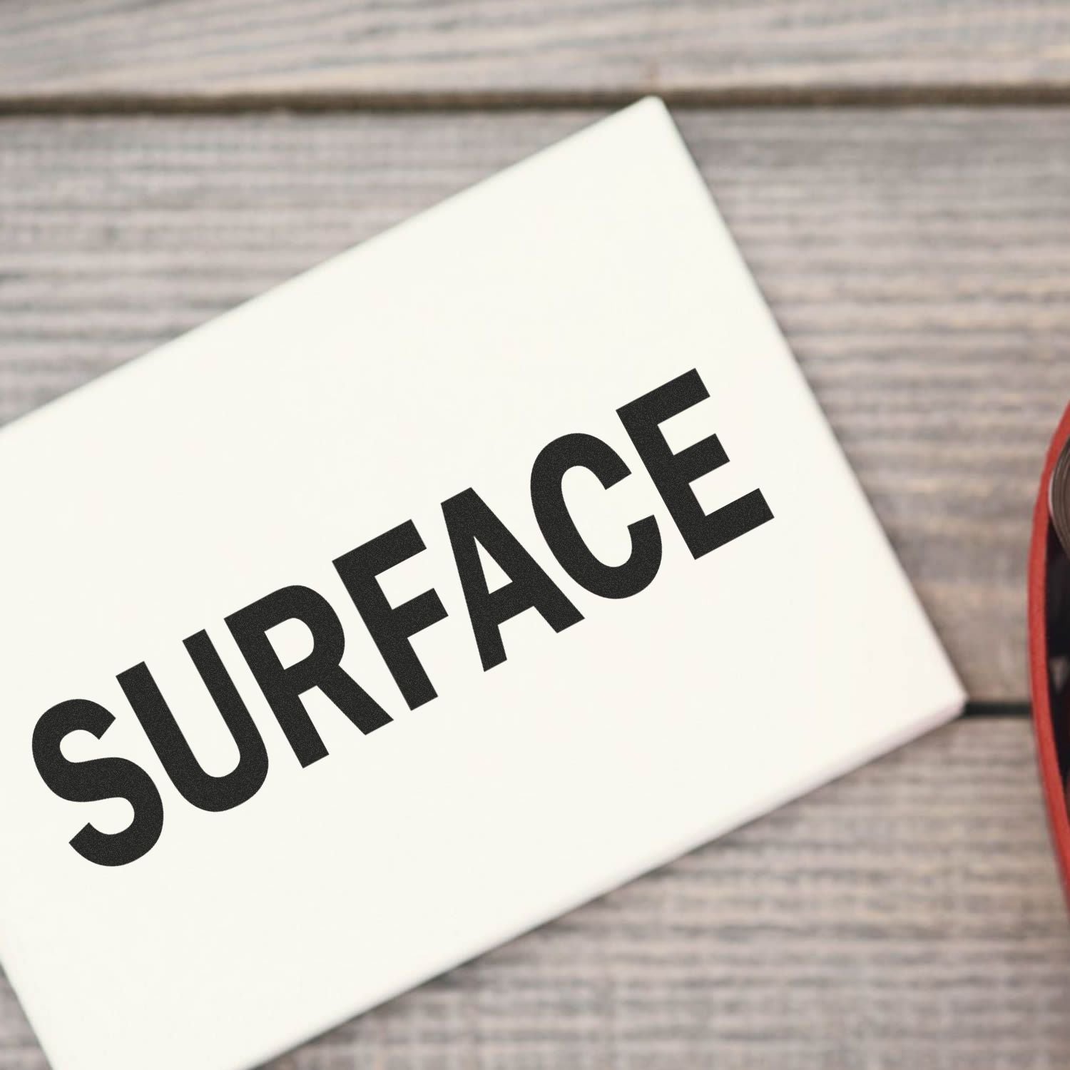 Self Inking Surface Stamp in use, displaying the word SURFACE on a white card placed on a wooden surface.