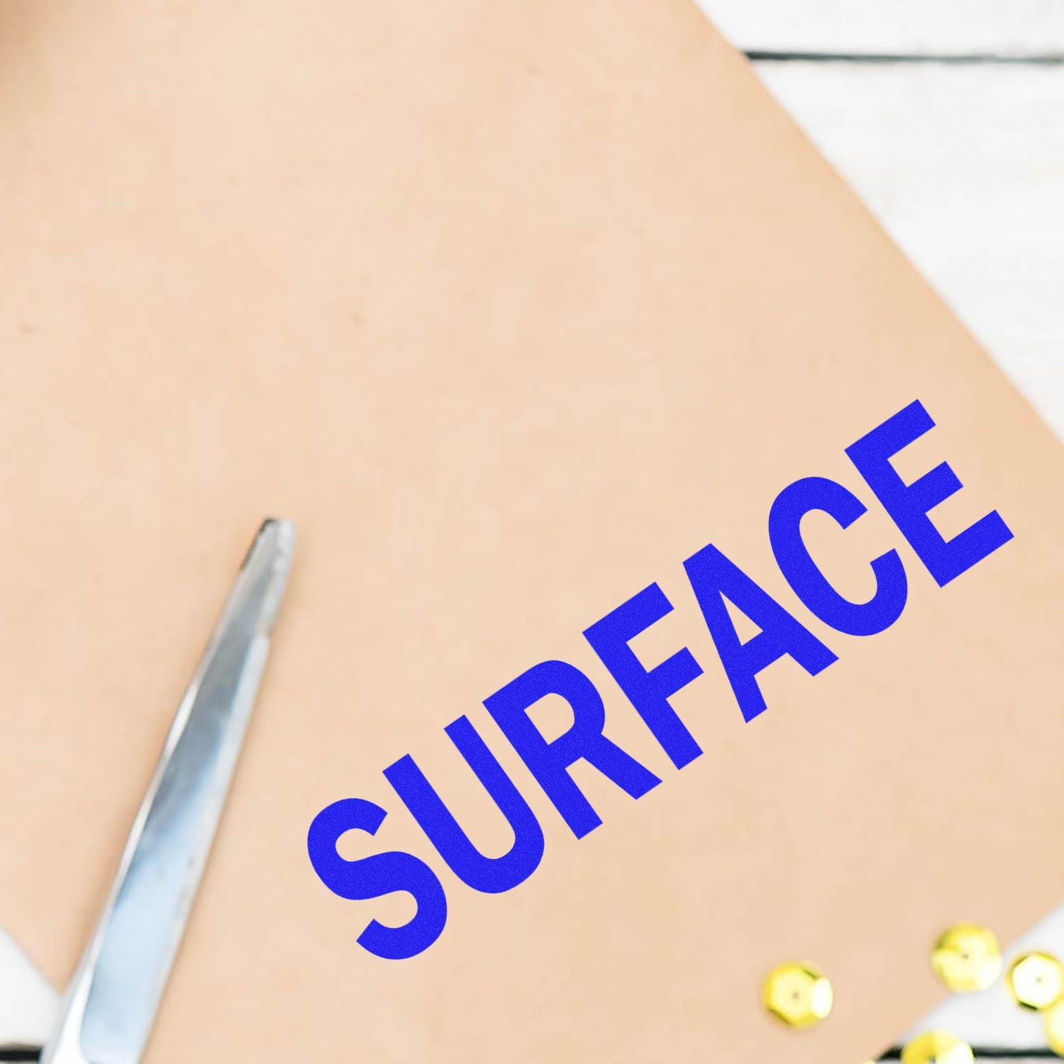 Large Surface Rubber Stamp imprinting the word SURFACE in blue on a beige sheet, with a pair of scissors and sequins nearby.