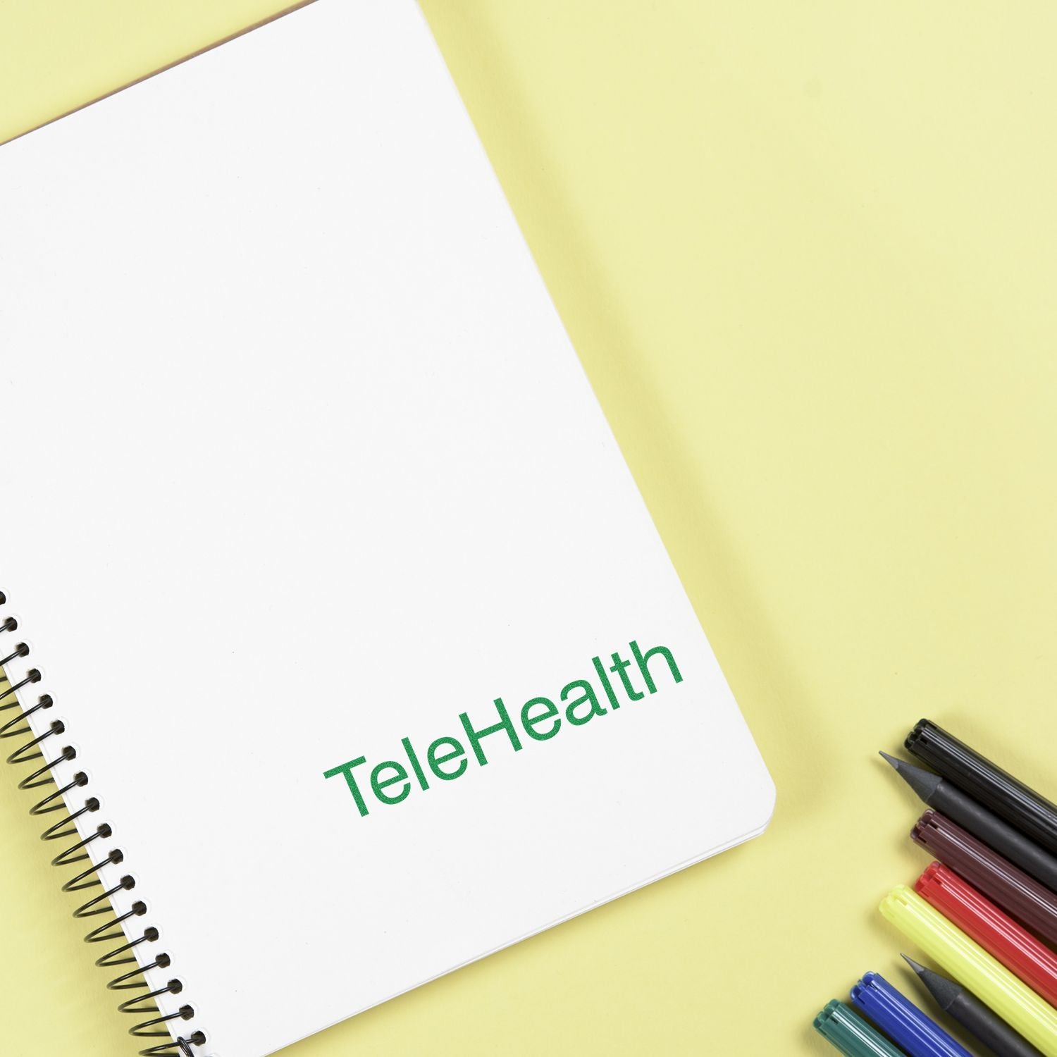 Large TeleHealth rubber stamp on a white notebook with spiral binding, placed on a yellow surface next to colorful pens.