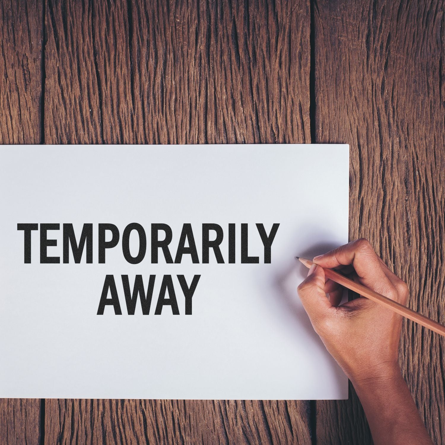 Hand holding a large temporarily away rubber stamp, pressing it onto a white paper with the text TEMPORARILY AWAY on a wooden surface.