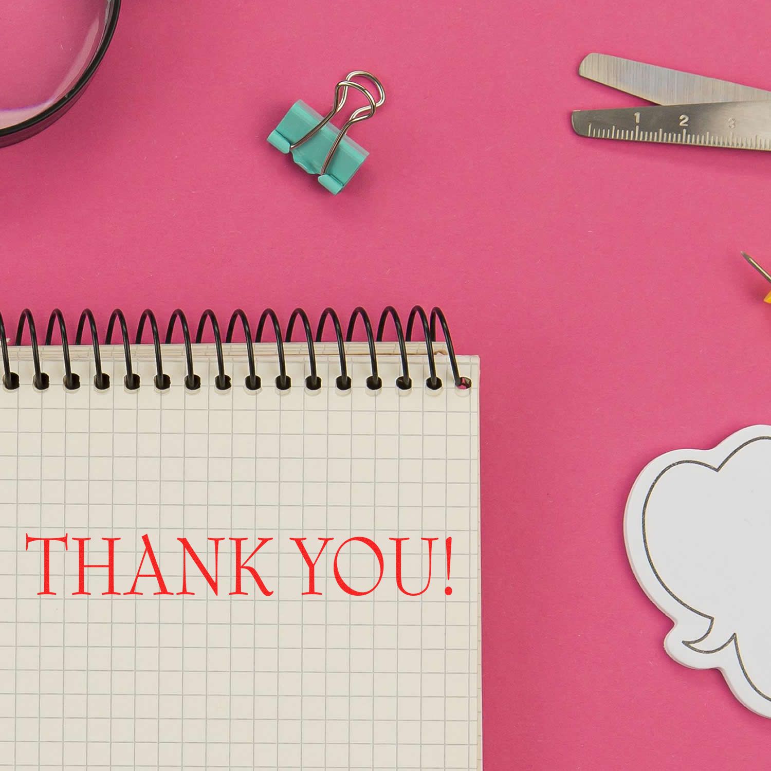 Large Self Inking Thank You Stamp in red ink on a spiral notebook, surrounded by office supplies on a pink background.