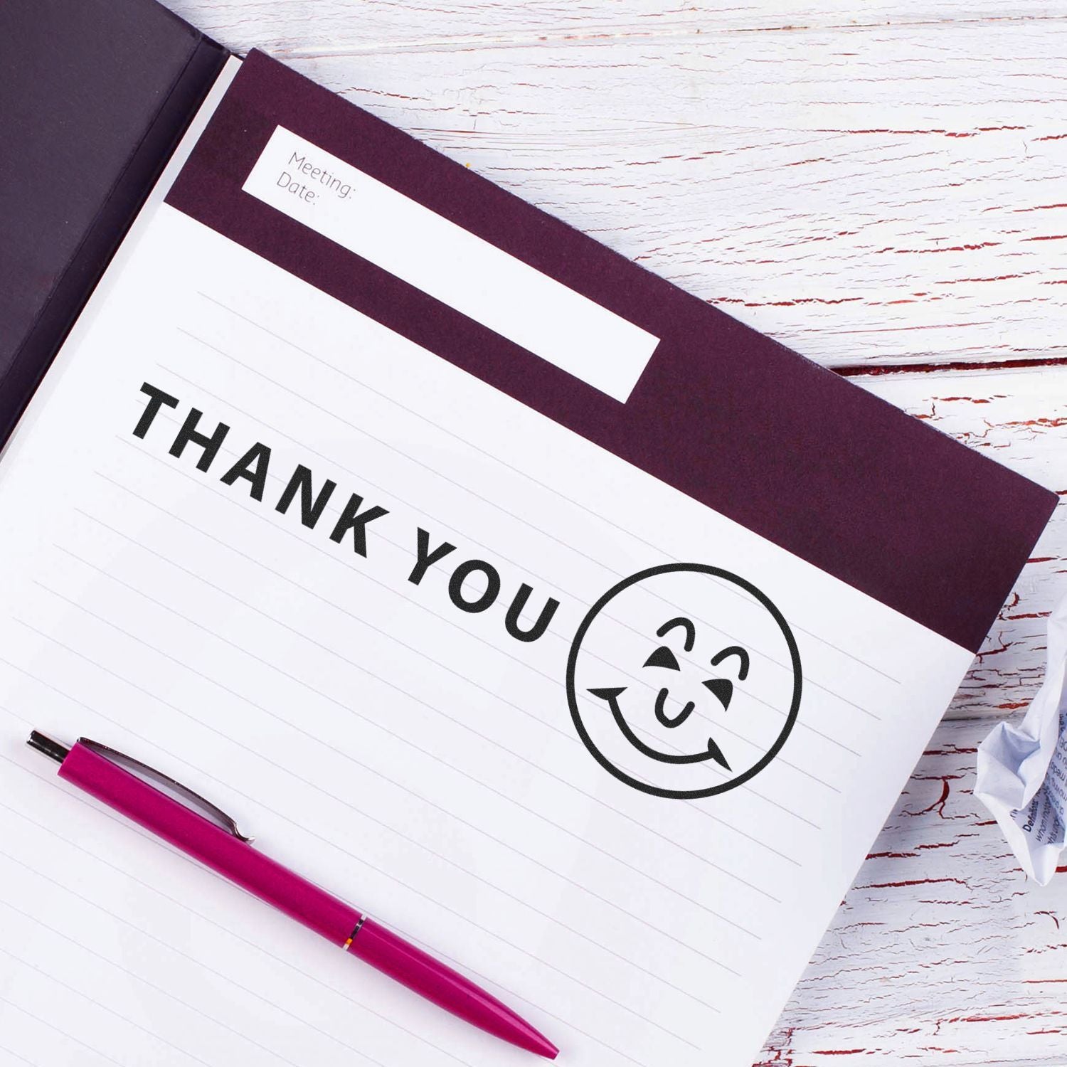 Thank You with Smiley Rubber Stamp on a notebook page with a pink pen, crumpled paper, and a black folder on a white wooden table.