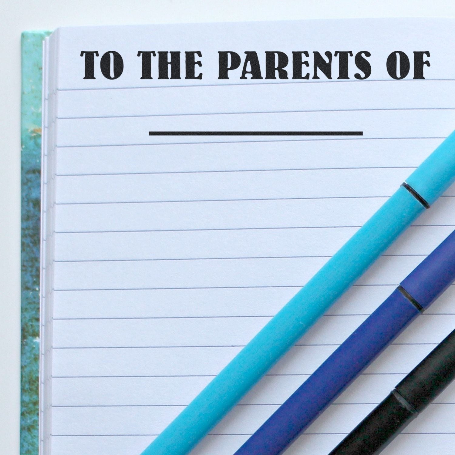 Notebook page with 'To The Parents Of' rubber stamp text at the top, and three pens placed diagonally on the right side.