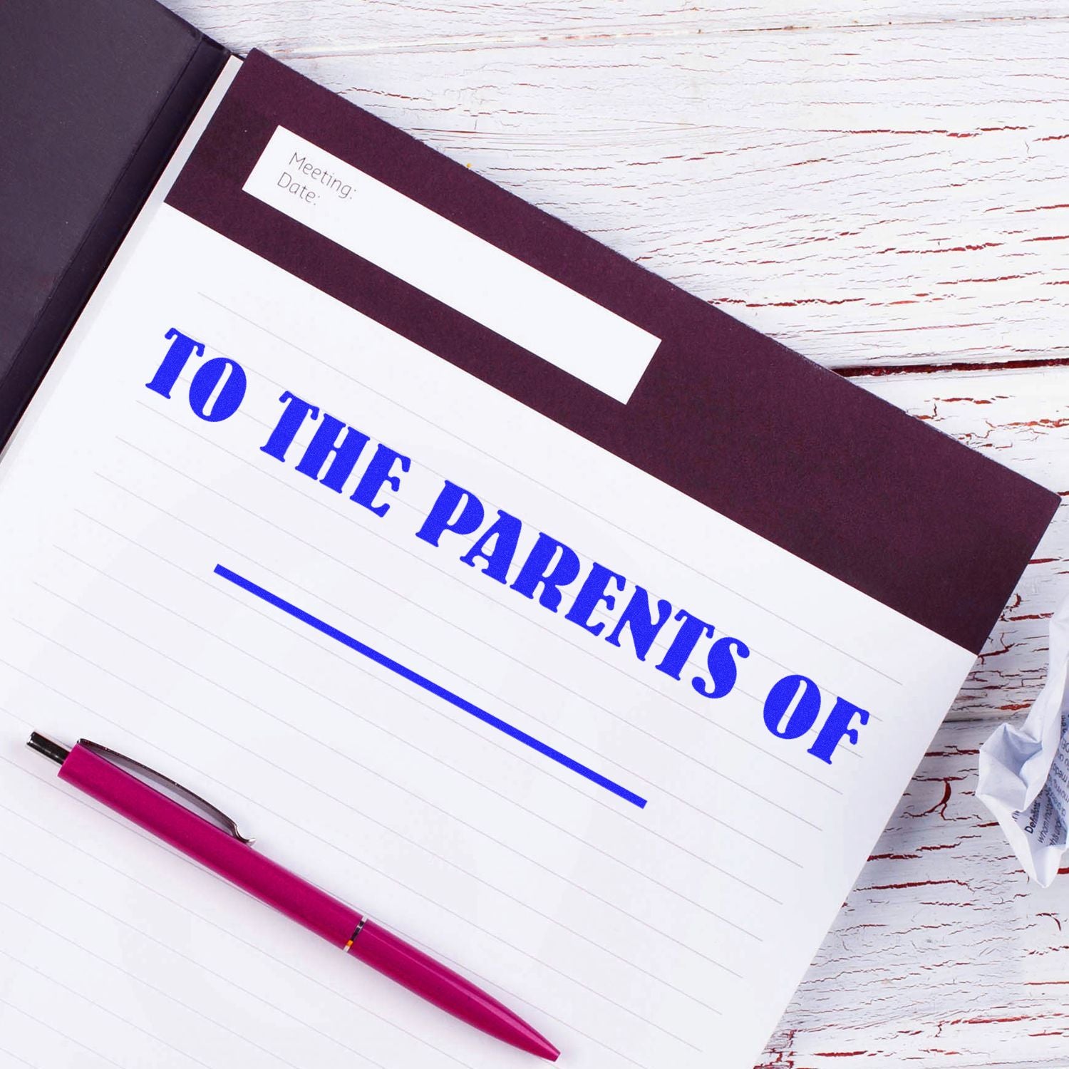 Notebook with 'To The Parents Of' rubber stamp imprint, pink pen, and crumpled paper on a white wooden table.