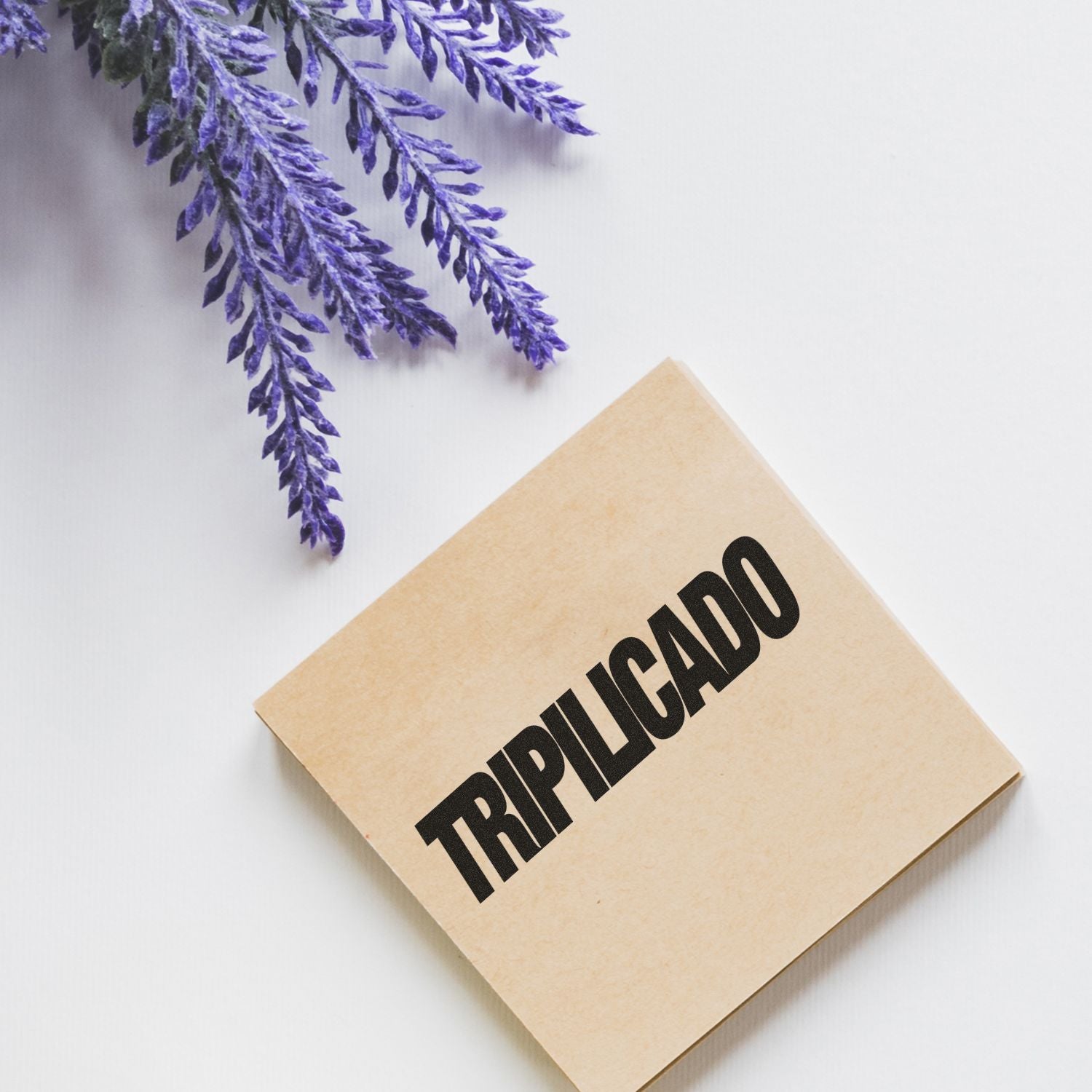 Large Pre-Inked Tripilicado Stamp in use, stamping TRIPLICADO on a brown paper, with purple flowers in the background.