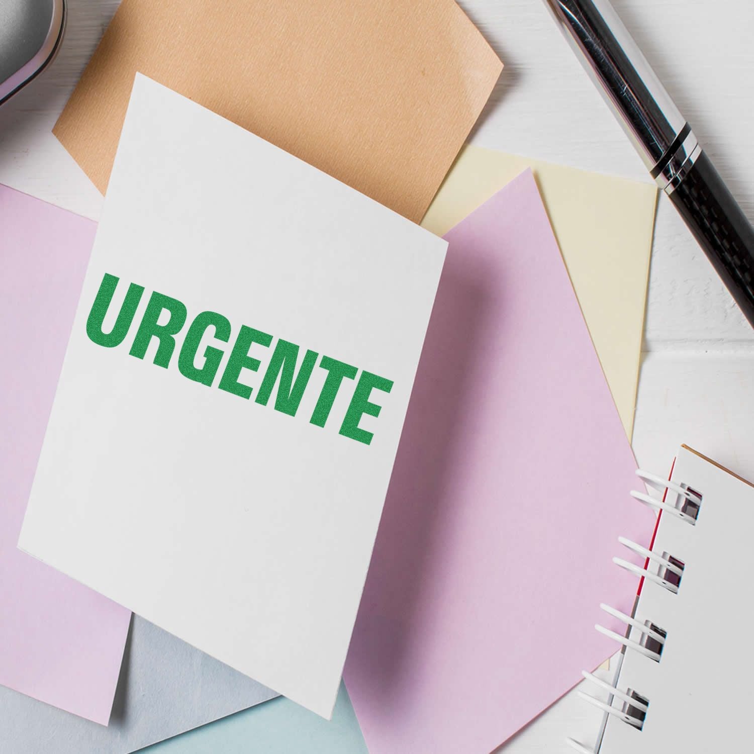 Large Urgente rubber stamp on white paper surrounded by pastel-colored sheets, a pen, and a notebook.