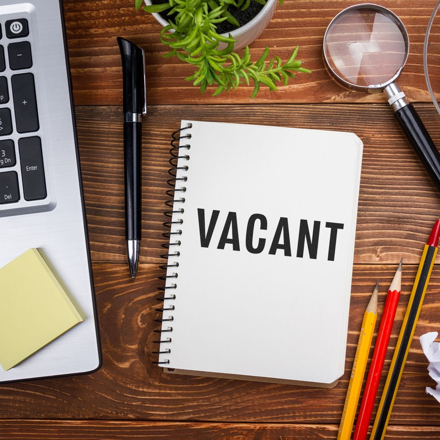 Large vacant rubber stamp on a spiral notebook, surrounded by office supplies including a pen, magnifying glass, and plant.