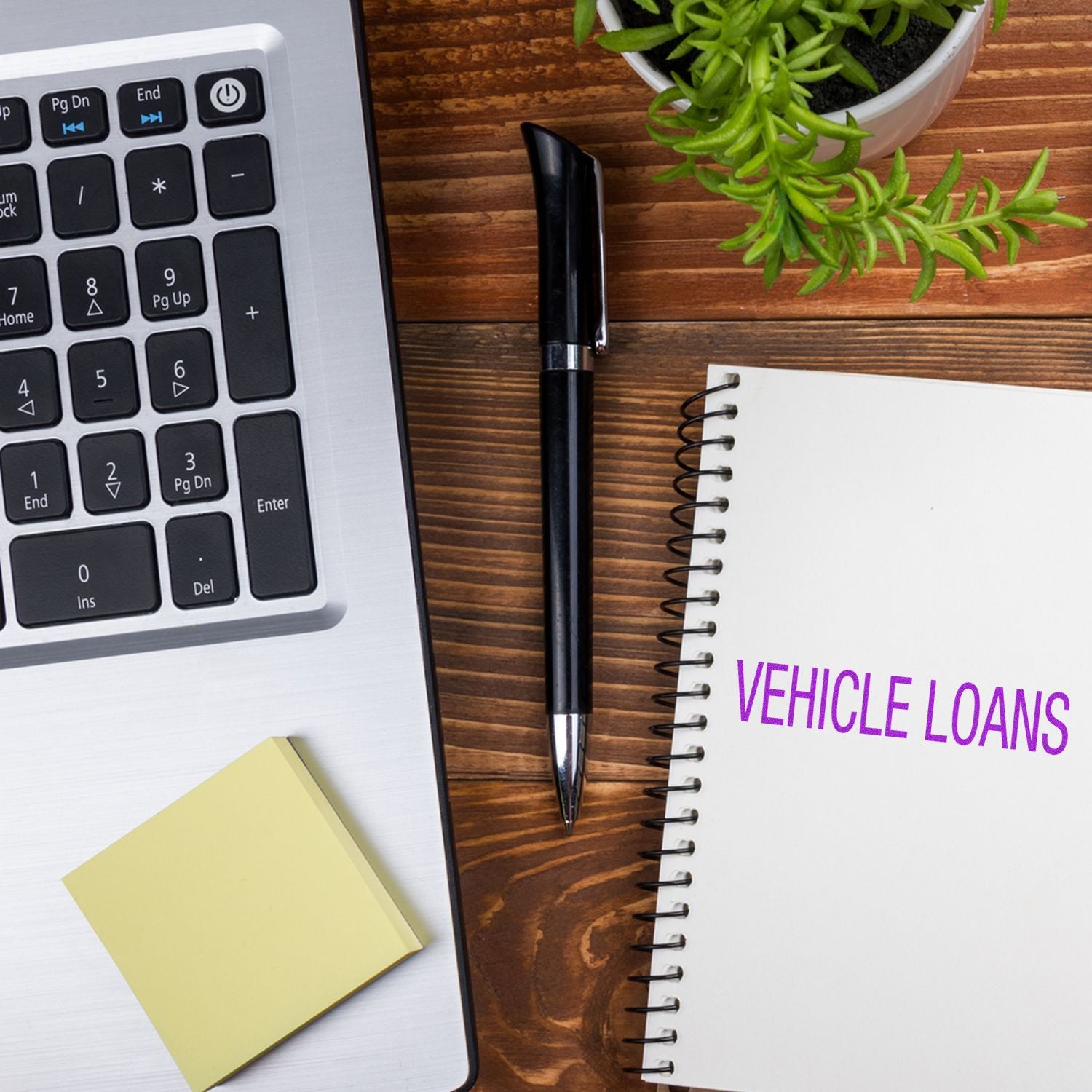 Laptop, pen, sticky notes, plant, and notebook with Vehicle Loans stamped in purple on a wooden desk.