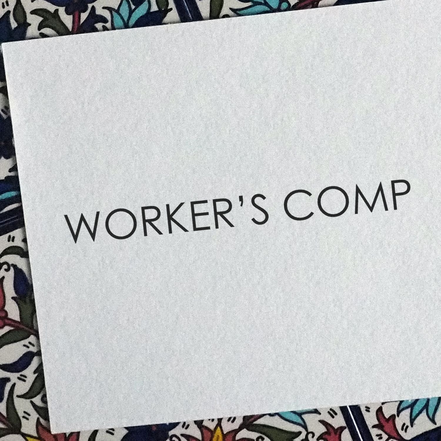 Slim Pre-Inked Workers Comp Stamp imprint on white paper with WORKER'S COMP text, placed on a colorful patterned surface.