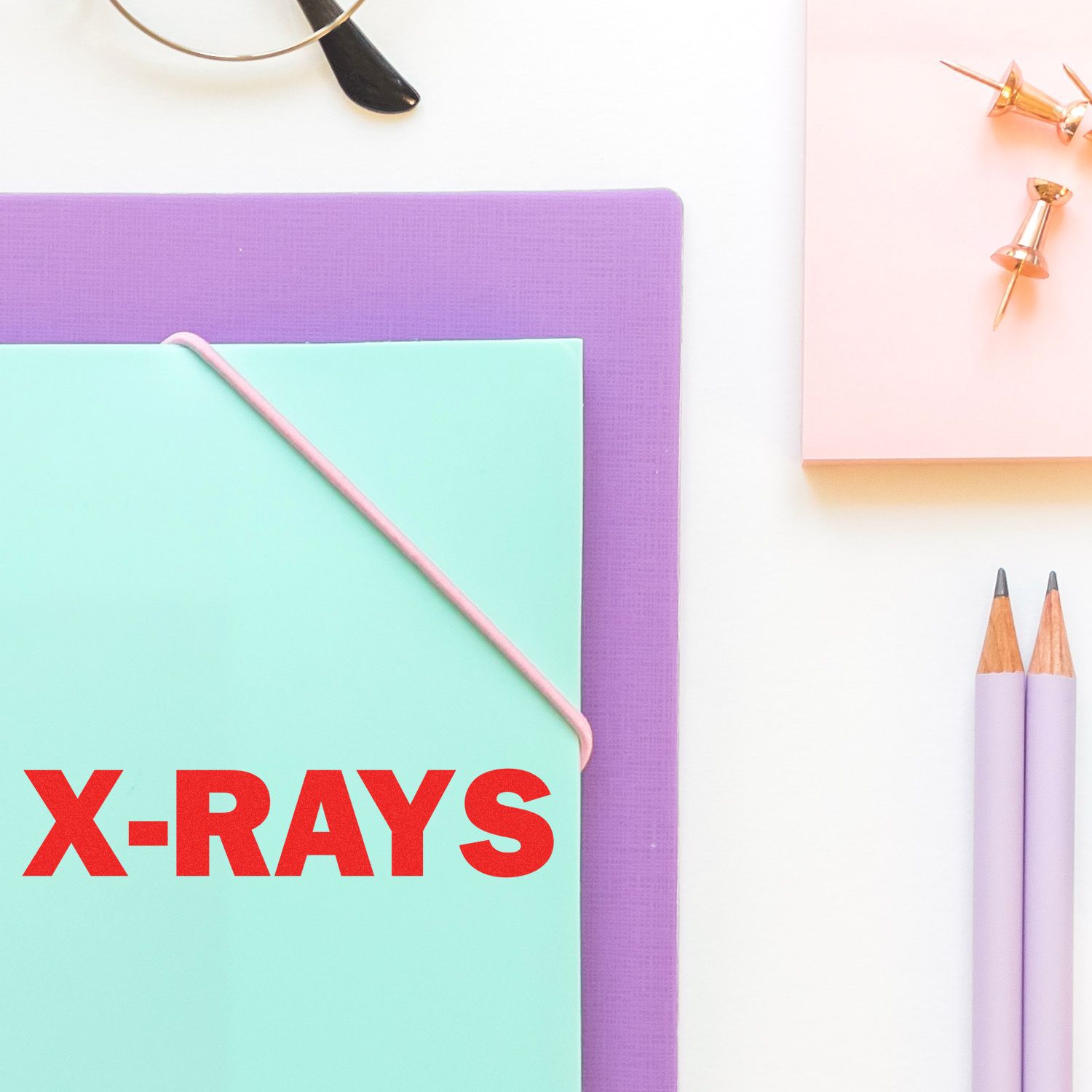 Large X-Rays rubber stamp in red ink on a mint green folder, surrounded by office supplies including pencils, push pins, and glasses.