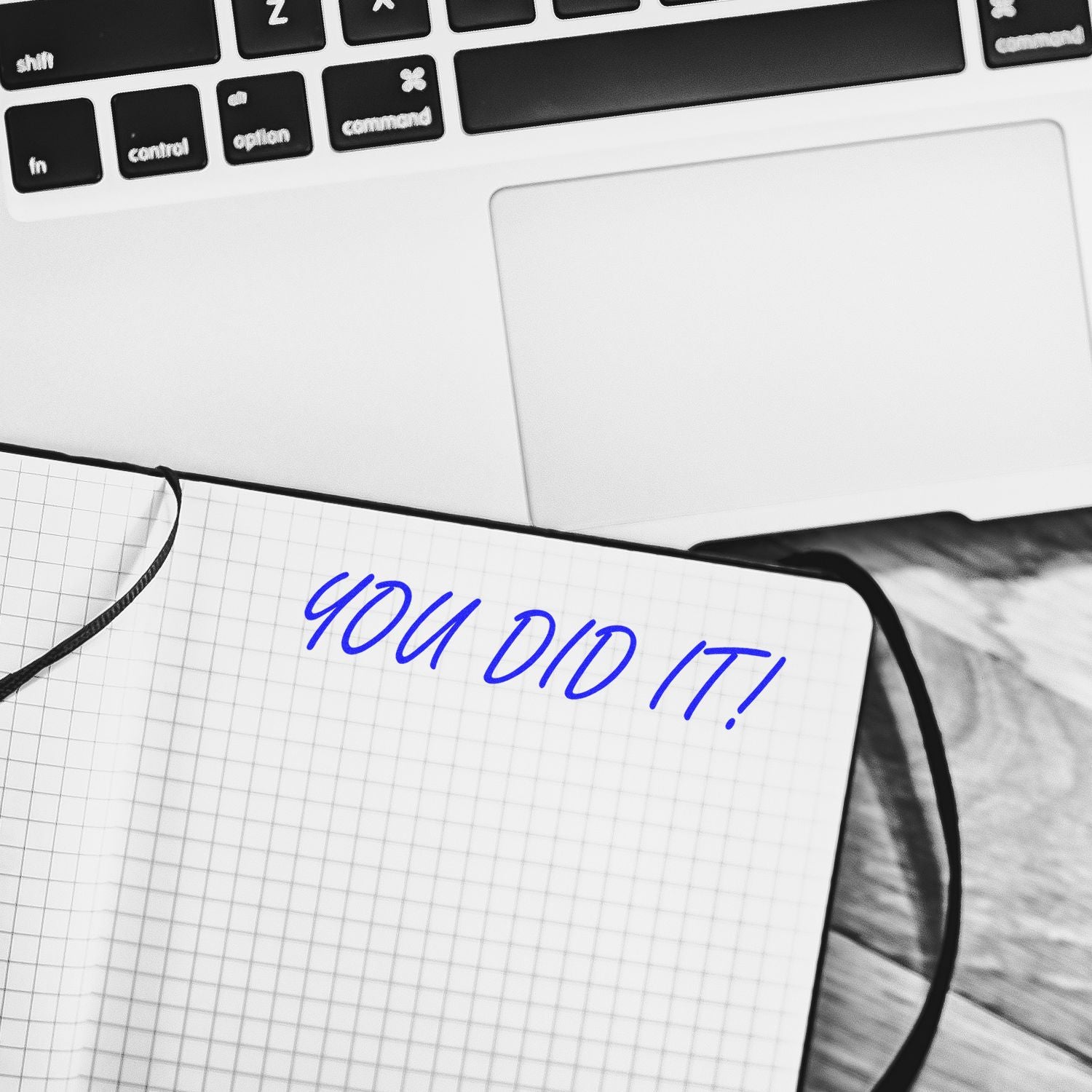 A Self Inking You Did It Stamp imprint in blue ink on a notebook, placed next to a laptop on a wooden desk.