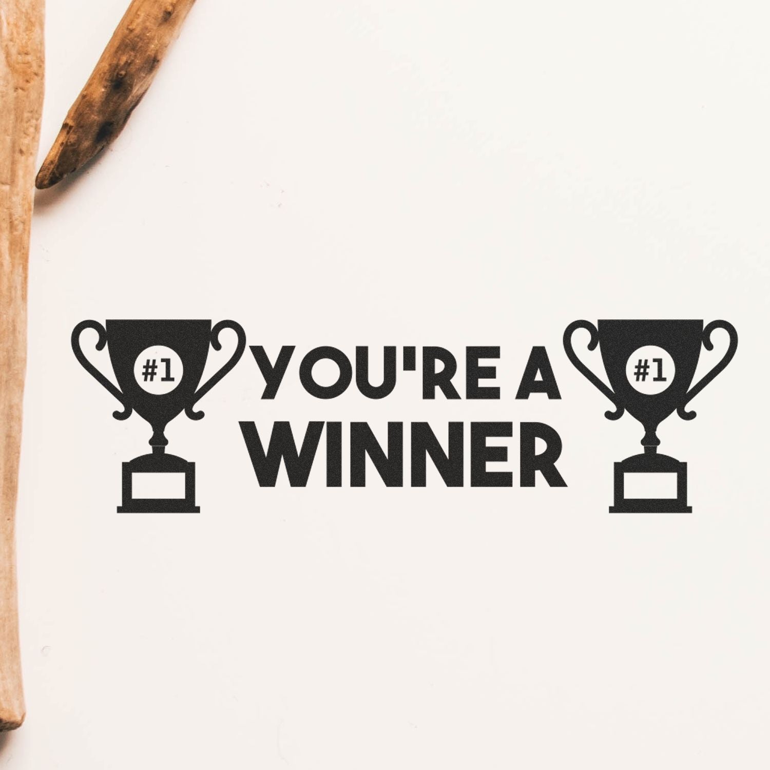 Large You're a Winner rubber stamp impression with two trophy icons on a white background, next to a wooden stick.