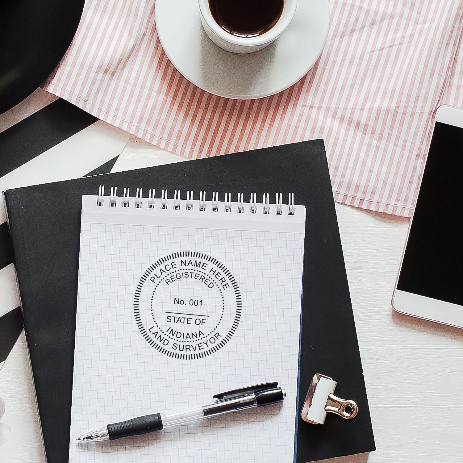 Self Inking Indiana Land Surveyor Stamp on a notepad with a pen, clipboard, coffee cup, and smartphone on a desk.