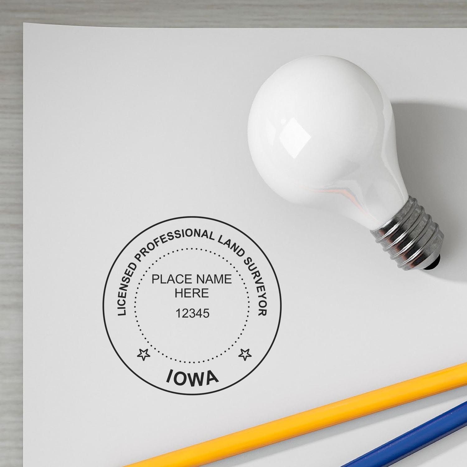 Self Inking Iowa Land Surveyor Stamp on white paper with a light bulb and pencils nearby.