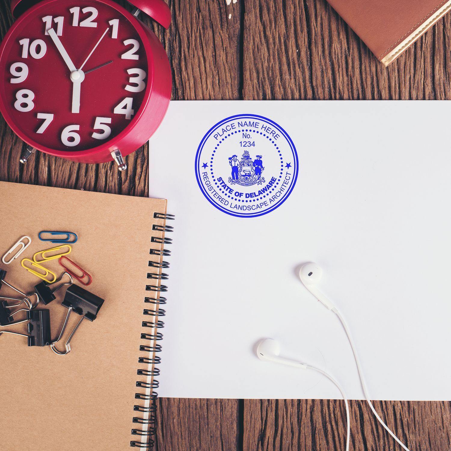 A desk with a red clock, notebook, paper clips, and earphones. The Landscape Architect Regular Rubber Stamp of Seal is stamped on white paper.