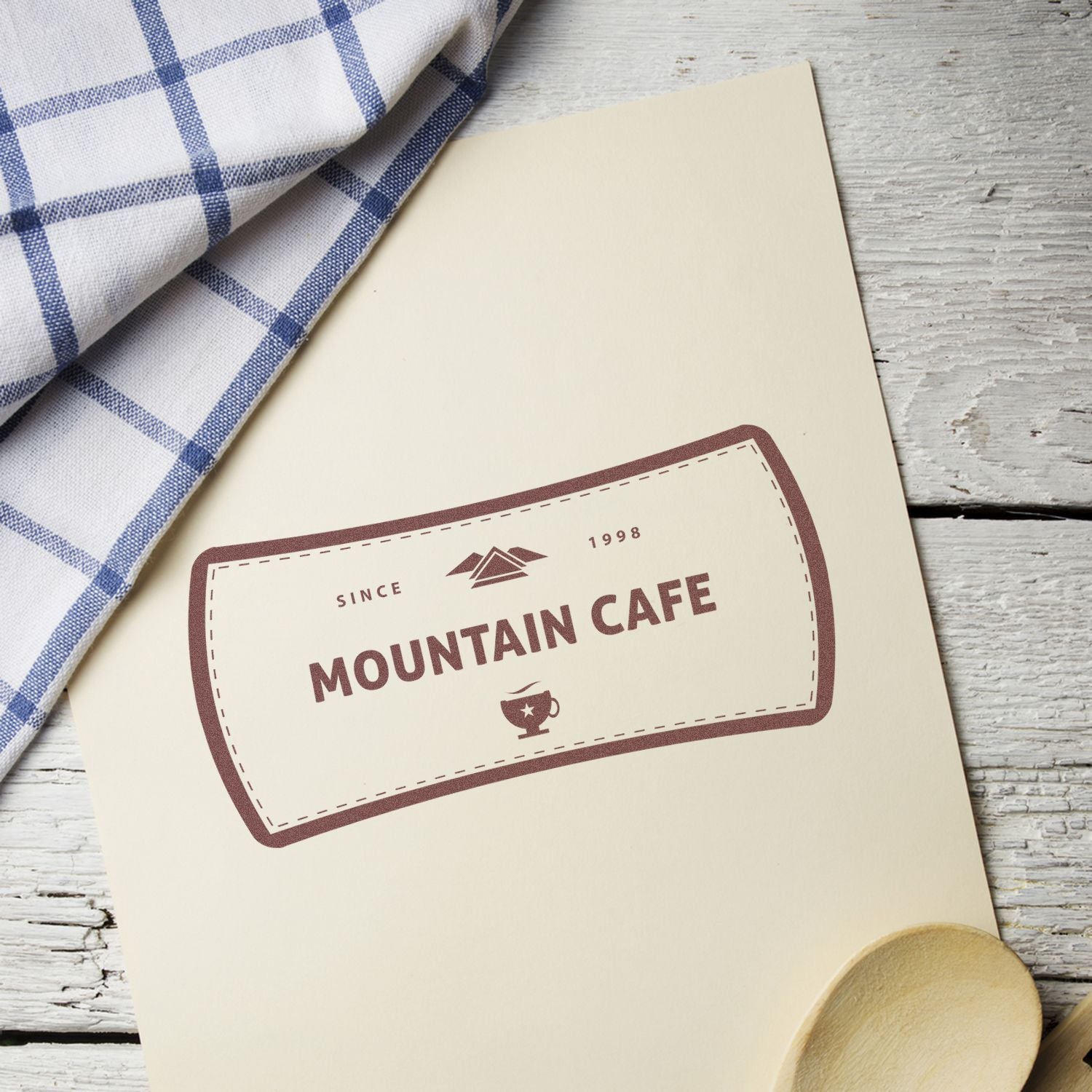 Custom Rubber Stamp Size 2 x 5 used to stamp Mountain Cafe logo on a beige paper, placed on a rustic wooden table with a cloth and spoon.