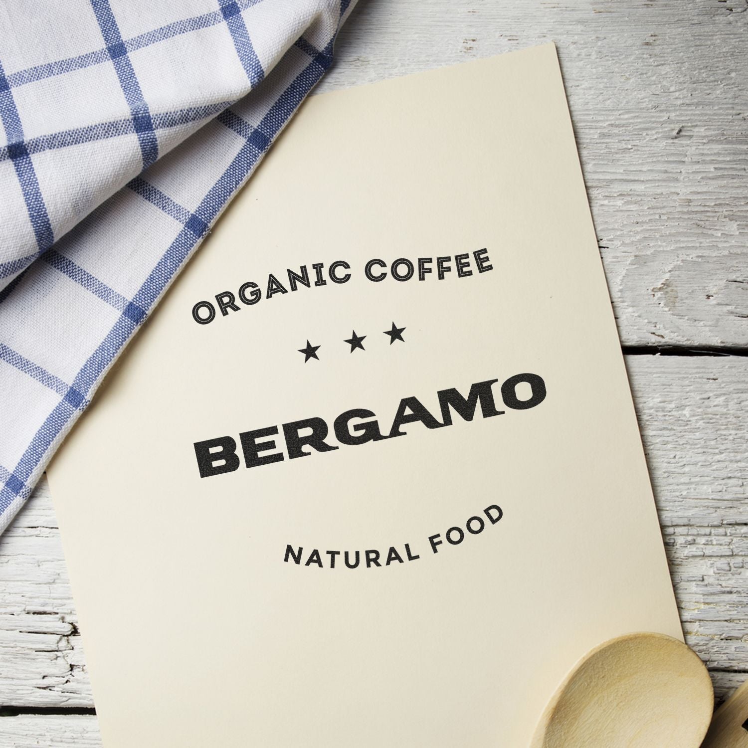 Custom Rubber Stamp Size 4 x 7 used to stamp Organic Coffee Bergamo Natural Food on a beige paper, next to a checkered cloth and wooden spoon.