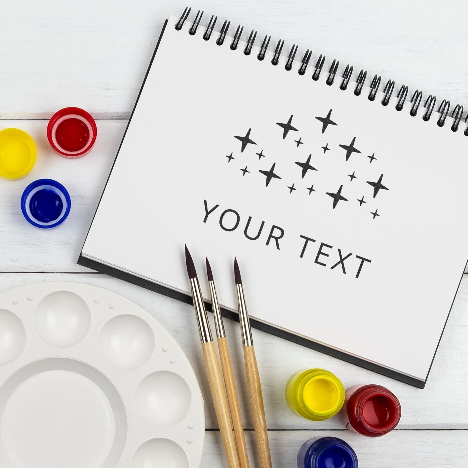 Custom Rubber Stamp Size 4 x 6 in use on a spiral notebook with paintbrushes, paint pots, and a palette on a white wooden surface.