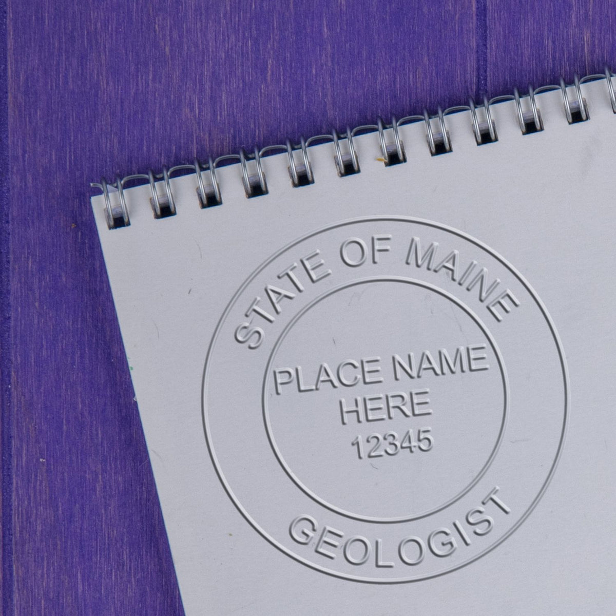 A lifestyle photo showing a stamped image of the Handheld Maine Professional Geologist Embosser on a piece of paper