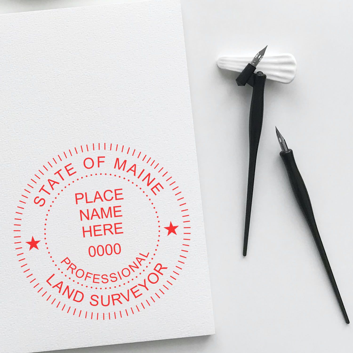 Premium MaxLight Pre-Inked Maine Surveyors Stamp in red ink on white paper, with two black drafting pens beside it.