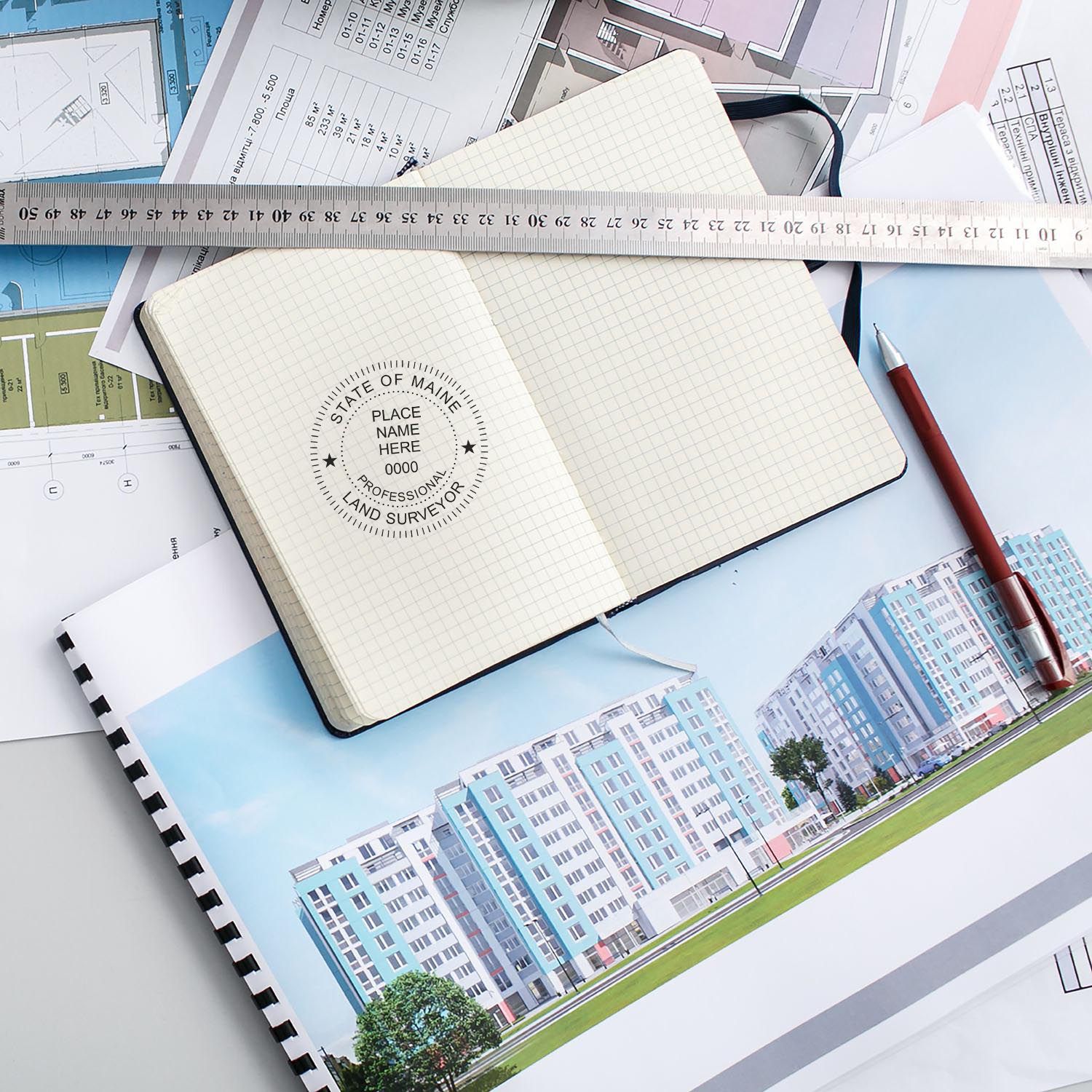 Self Inking Maine Land Surveyor Stamp in use on a notebook, surrounded by architectural plans, a ruler, and a red pen.