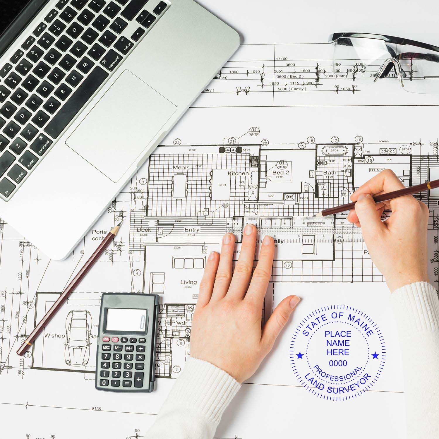 Person using Premium MaxLight Pre-Inked Maine Surveyors Stamp on architectural plans with a laptop, calculator, and glasses nearby.