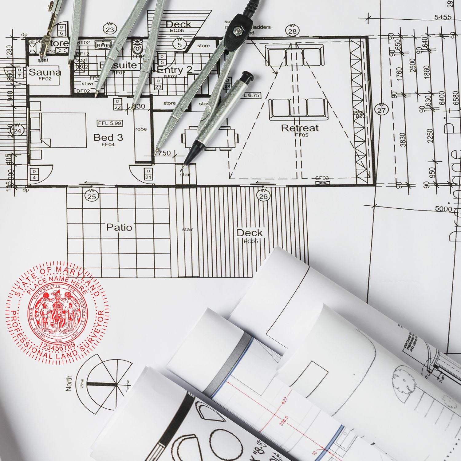 Blueprints with rolled plans, compass, and Premium MaxLight Pre-Inked Maryland Surveyors Stamp in red ink on architectural drawing.