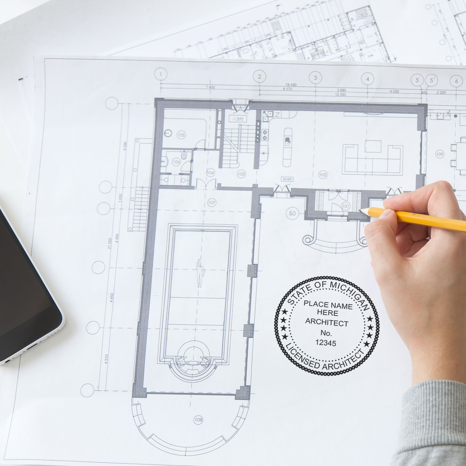 Hand holding a pencil over architectural blueprints with a Self Inking Michigan Architect Stamp imprint visible on the paper.