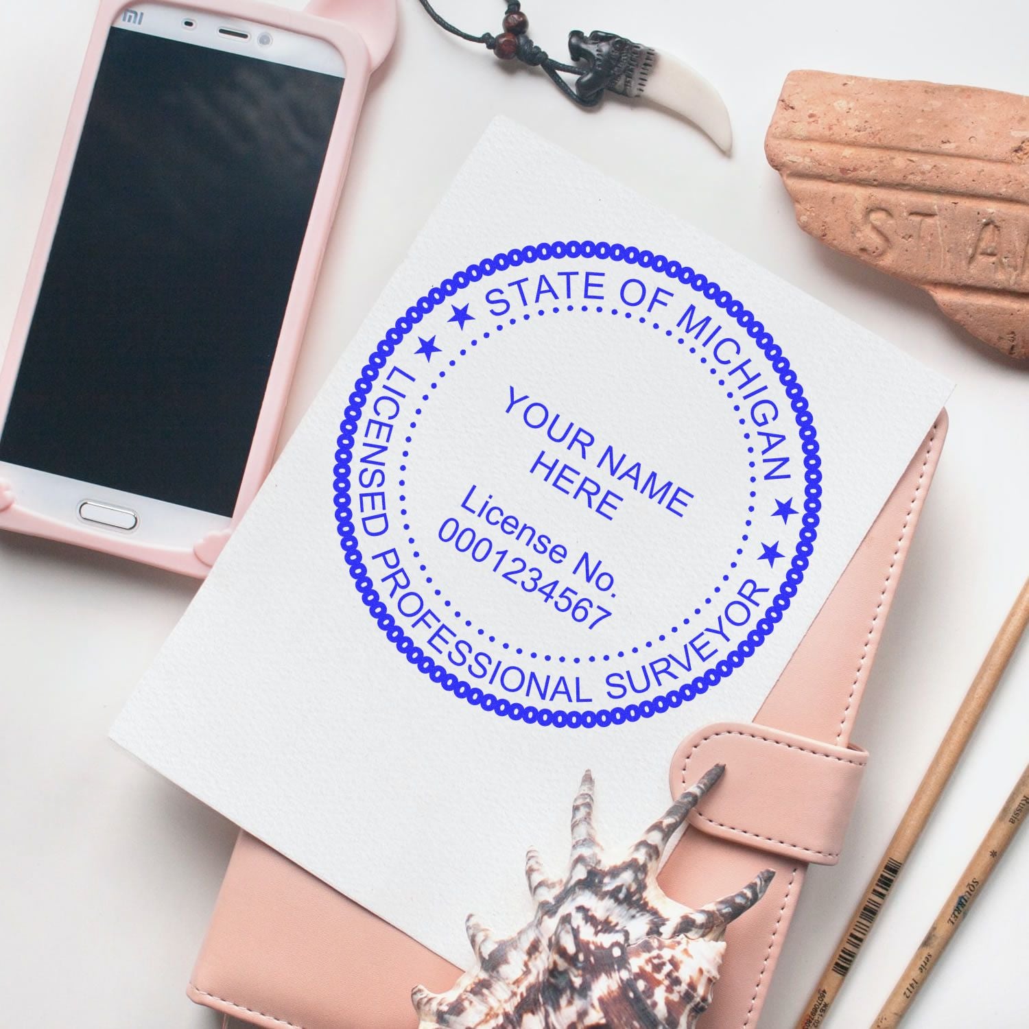 Self Inking Michigan Land Surveyor Stamp on white paper, surrounded by a phone, notebook, and decorative items.