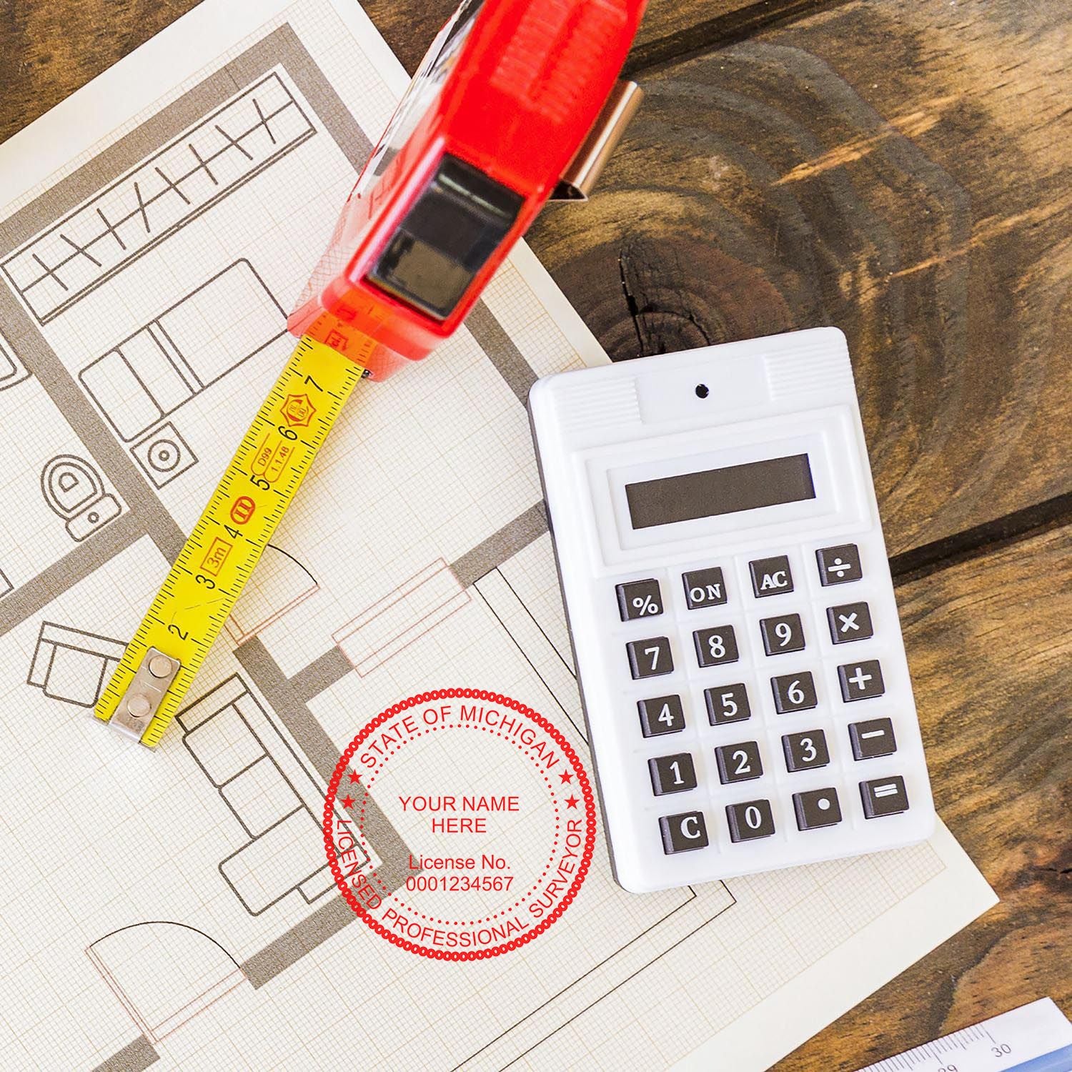 Self Inking Michigan Land Surveyor Stamp on architectural plans with a tape measure and calculator on a wooden surface.