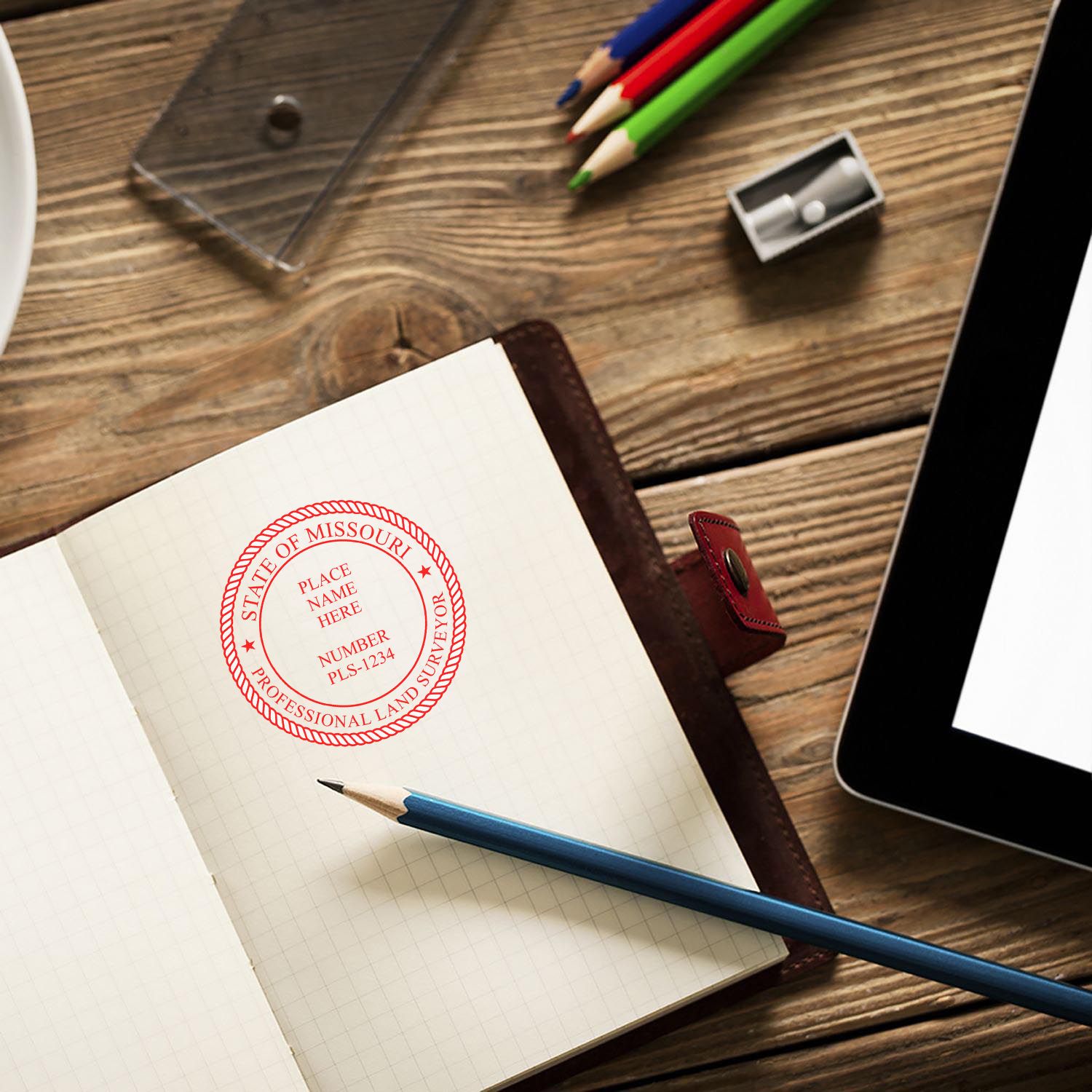 Self Inking Missouri Land Surveyor Stamp on an open notebook, surrounded by pencils, a sharpener, and a tablet on a wooden desk.