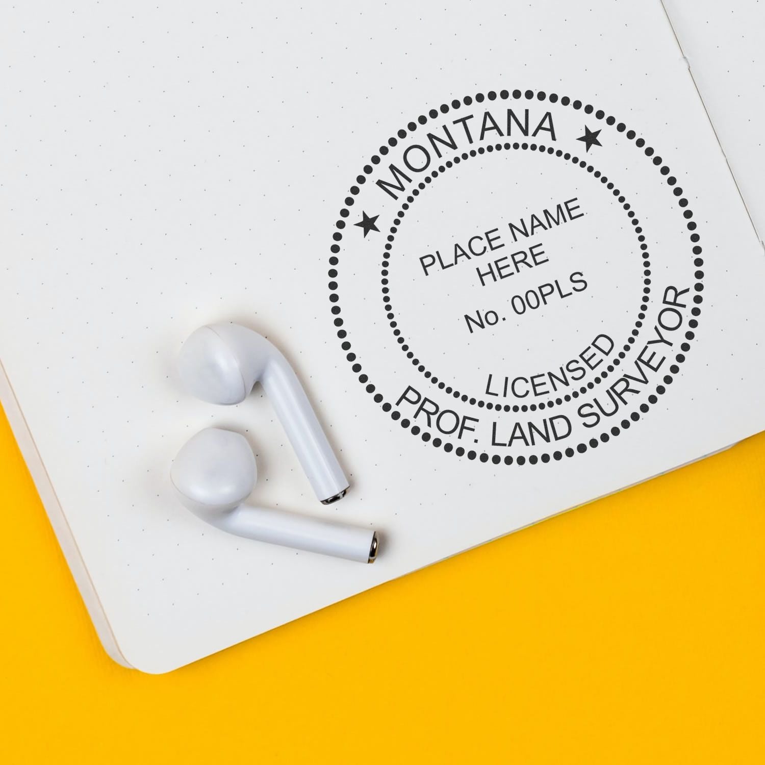 Montana Land Surveyor Seal Stamp, MT PLS Stamp, imprinted on a white notebook page with two white earbuds nearby on a yellow surface.