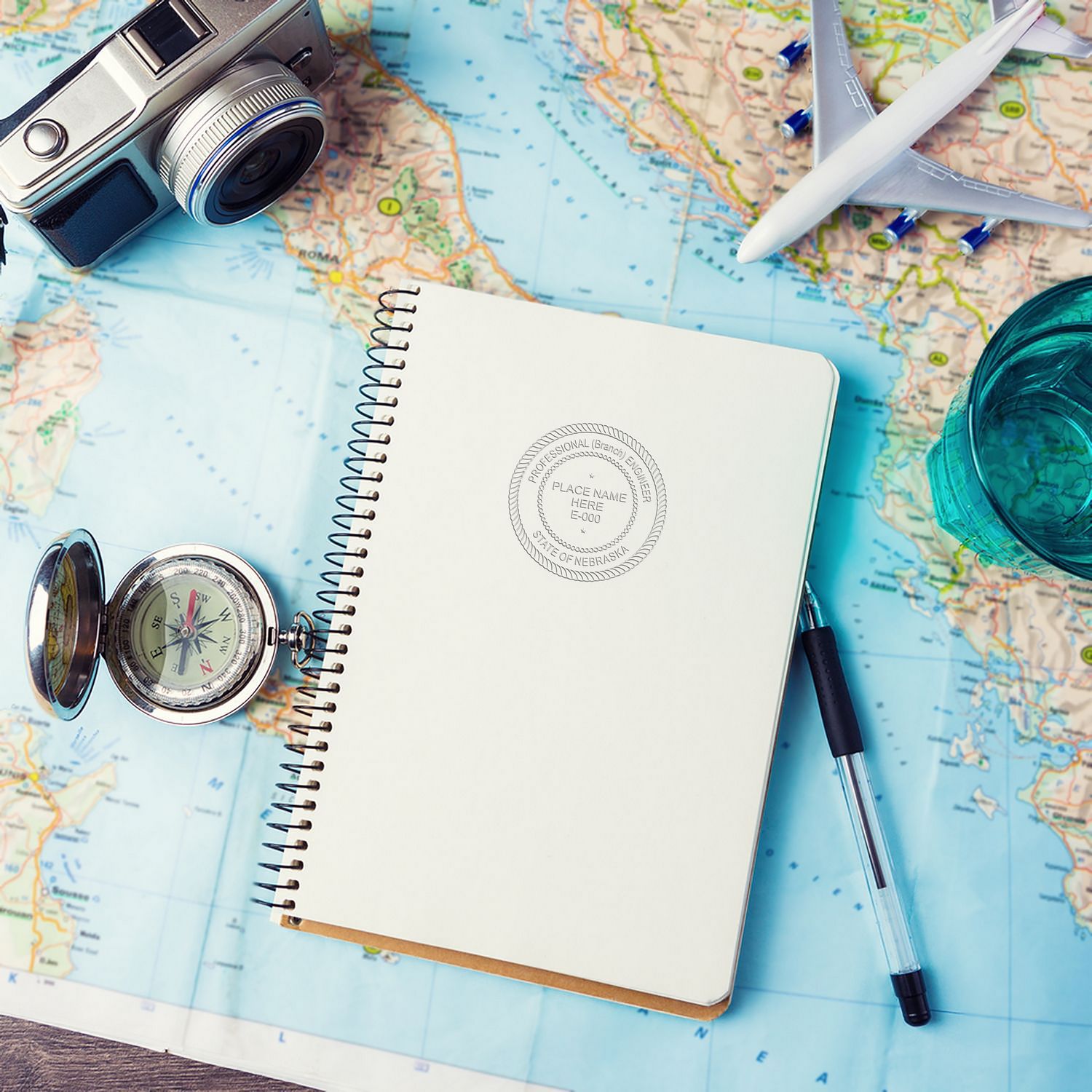 Architect Gold Gift Seal Embosser used on a notebook, placed on a world map with a camera, compass, pen, and airplane model nearby.