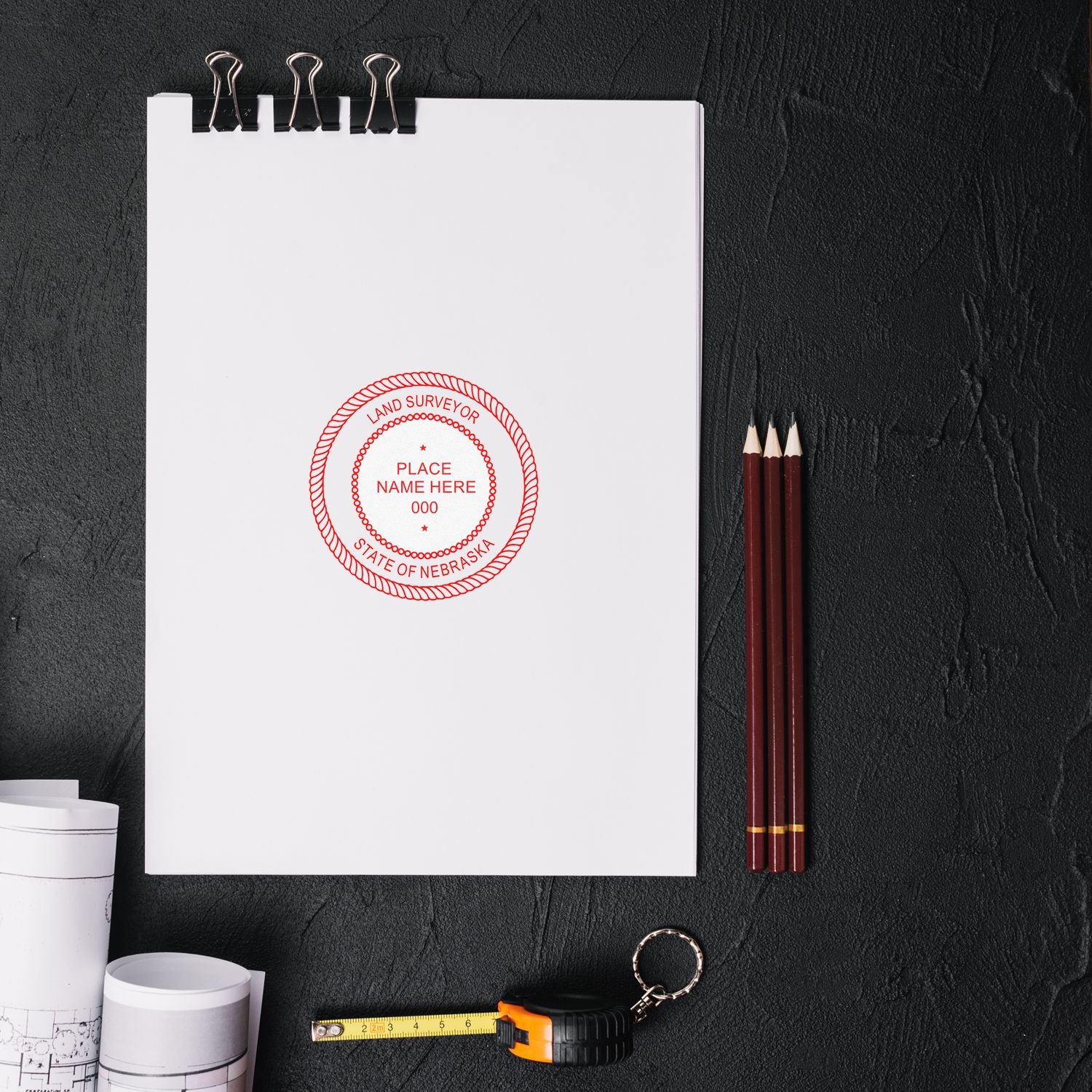 Self Inking Nebraska Land Surveyor Stamp on a white paper, surrounded by pencils, a tape measure, and rolled blueprints on a dark surface.