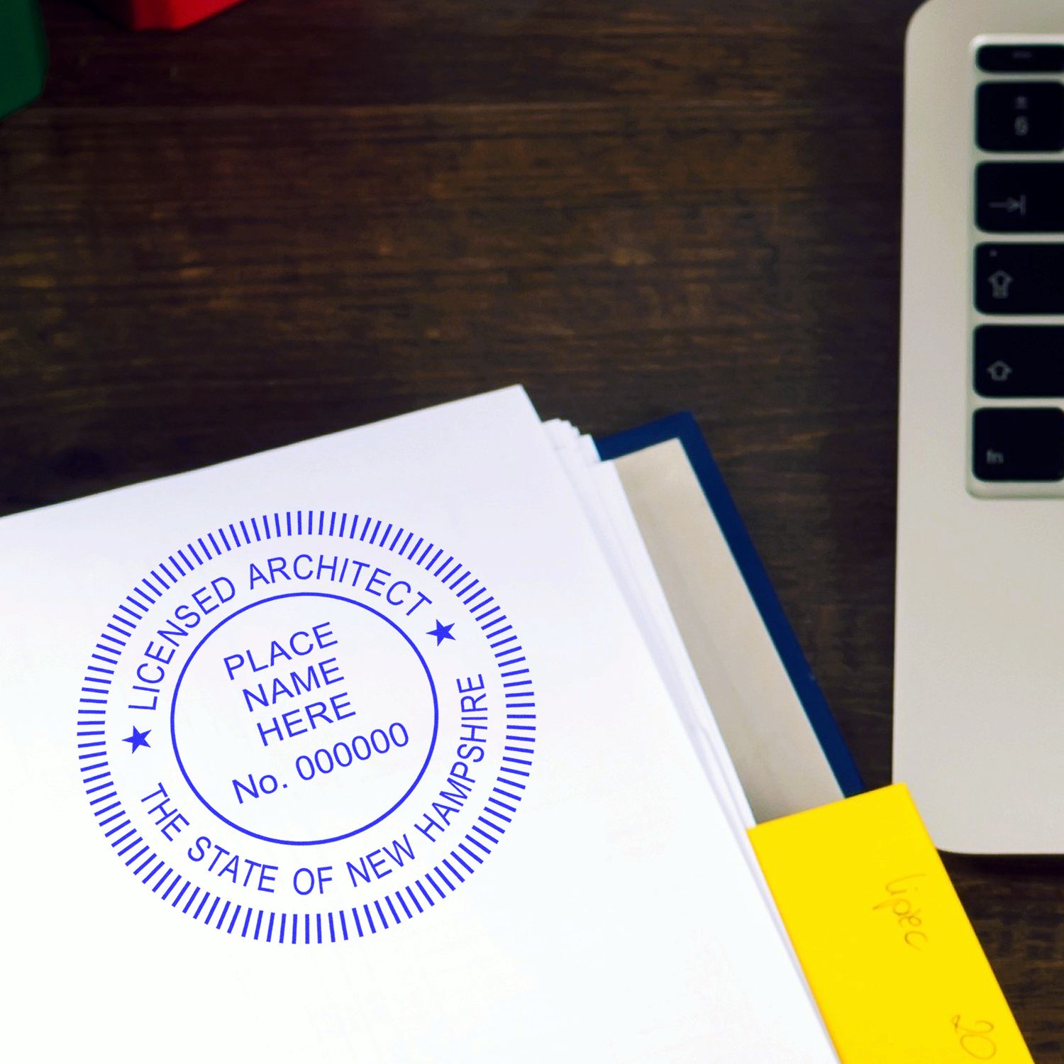 New Hampshire Architect Seal Stamp on a white document beside a laptop and a yellow sticky note on a wooden desk.