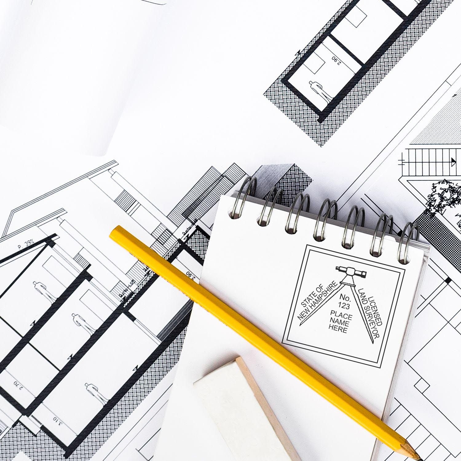 New Hampshire Land Surveyor Seal Stamp on a notepad with architectural blueprints, a yellow pencil, and an eraser.