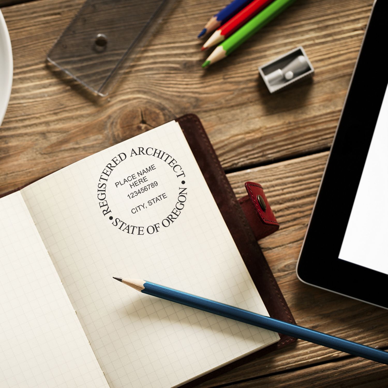 Premium MaxLight Pre-Inked Oregon Architectural Stamp in use on a notebook, surrounded by pencils, a sharpener, and a tablet on a wooden desk.