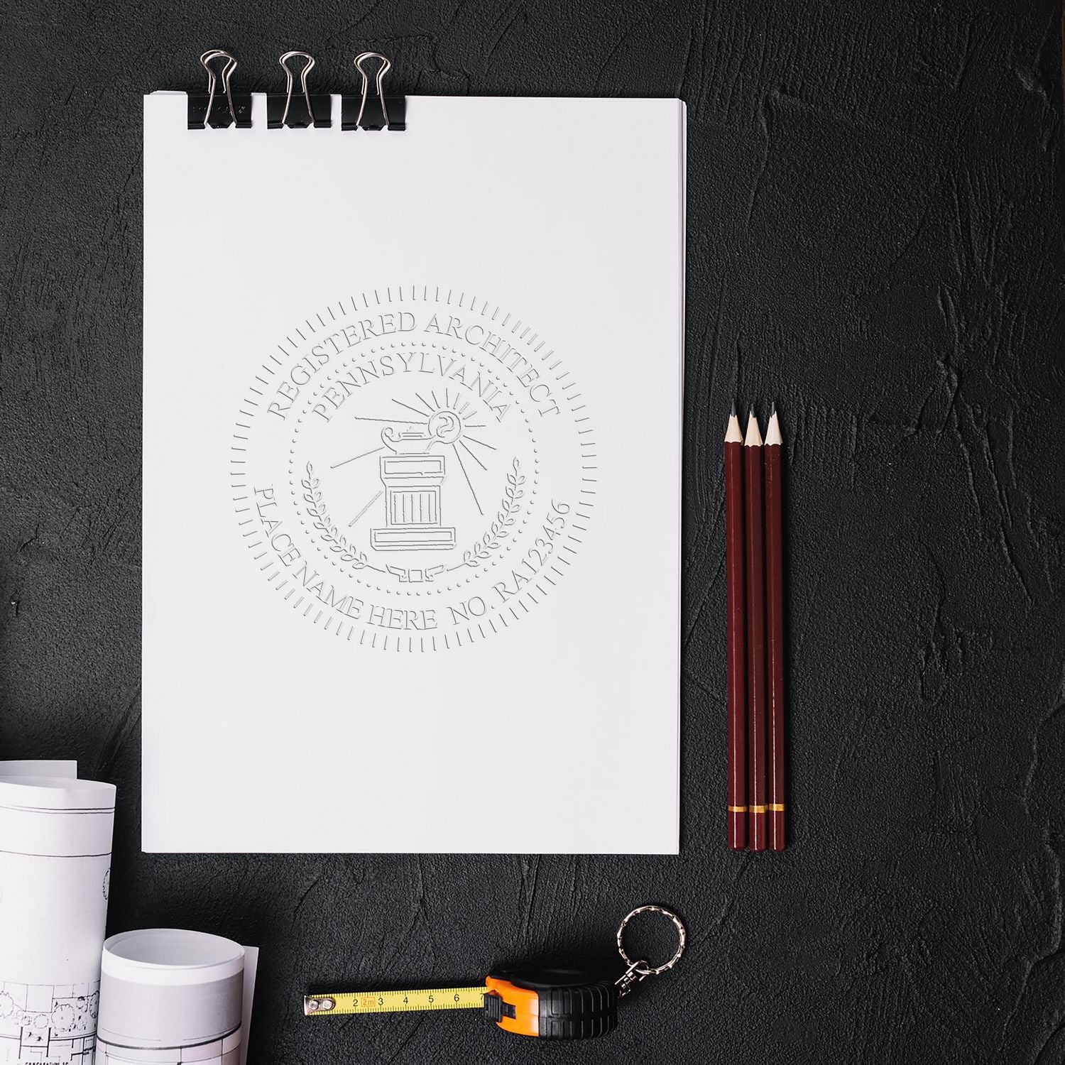 Architect Desk Seal Embosser imprint on a white sheet with architectural drawings, three pencils, a measuring tape, and a keychain on a black surface.