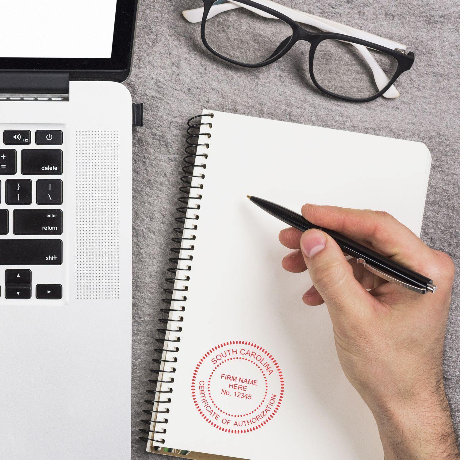 A hand holding a pen near a notebook stamped with the Professional Slim Pre-Inked Rubber Stamp of Seal, beside a laptop and glasses.