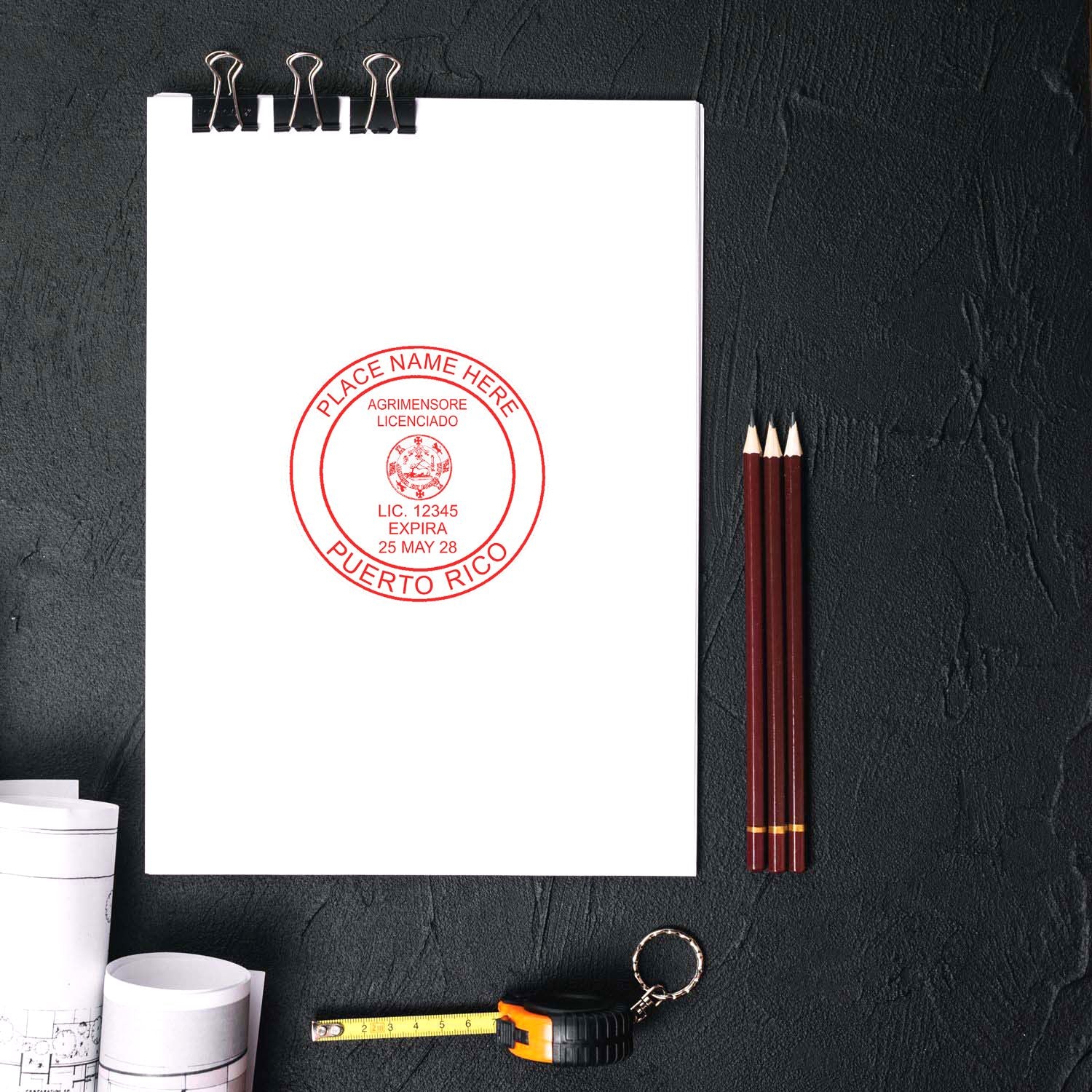 Puerto Rico Land Surveyor Seal Stamp on a white paper with pencils, a measuring tape, and blueprints on a dark surface.